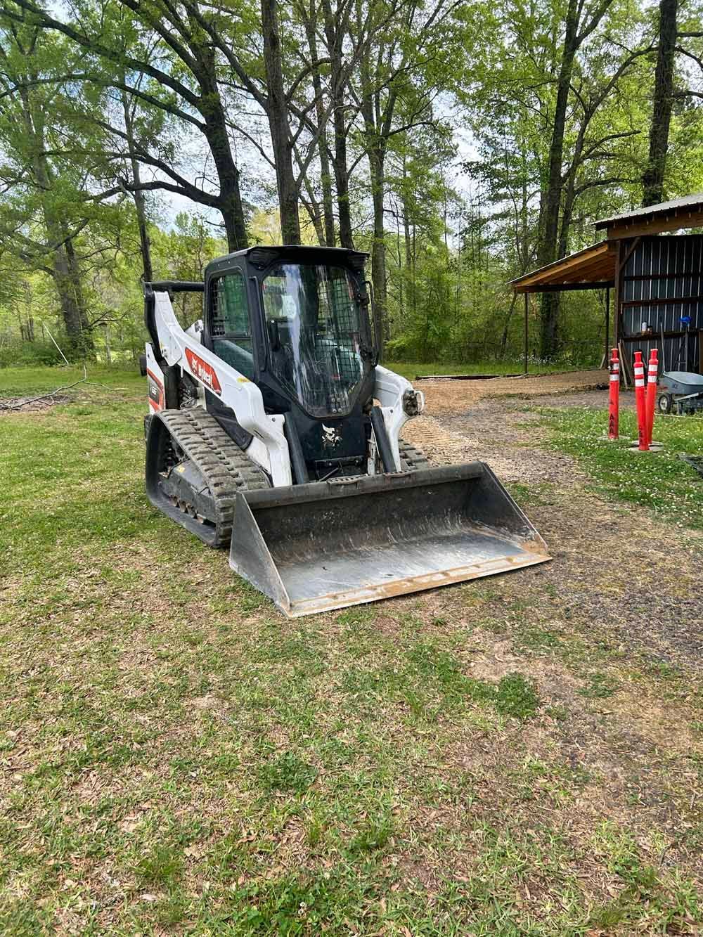 Compact loader parked on grass near trees, with front bucket lowered and a small shed nearby.