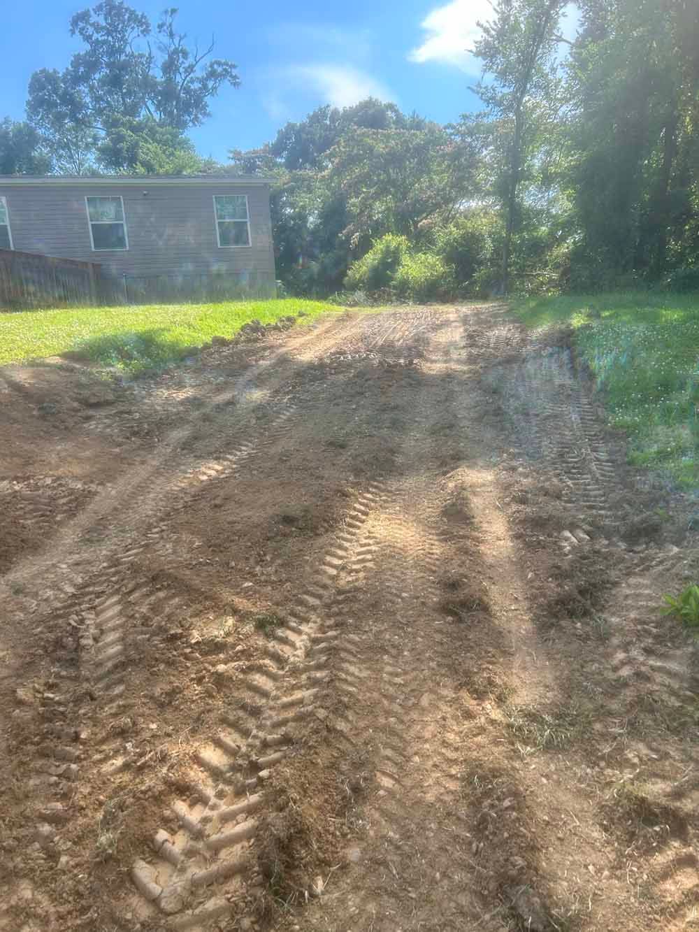 Freshly graded dirt driveway beside a house, with tire tracks and trees in the background