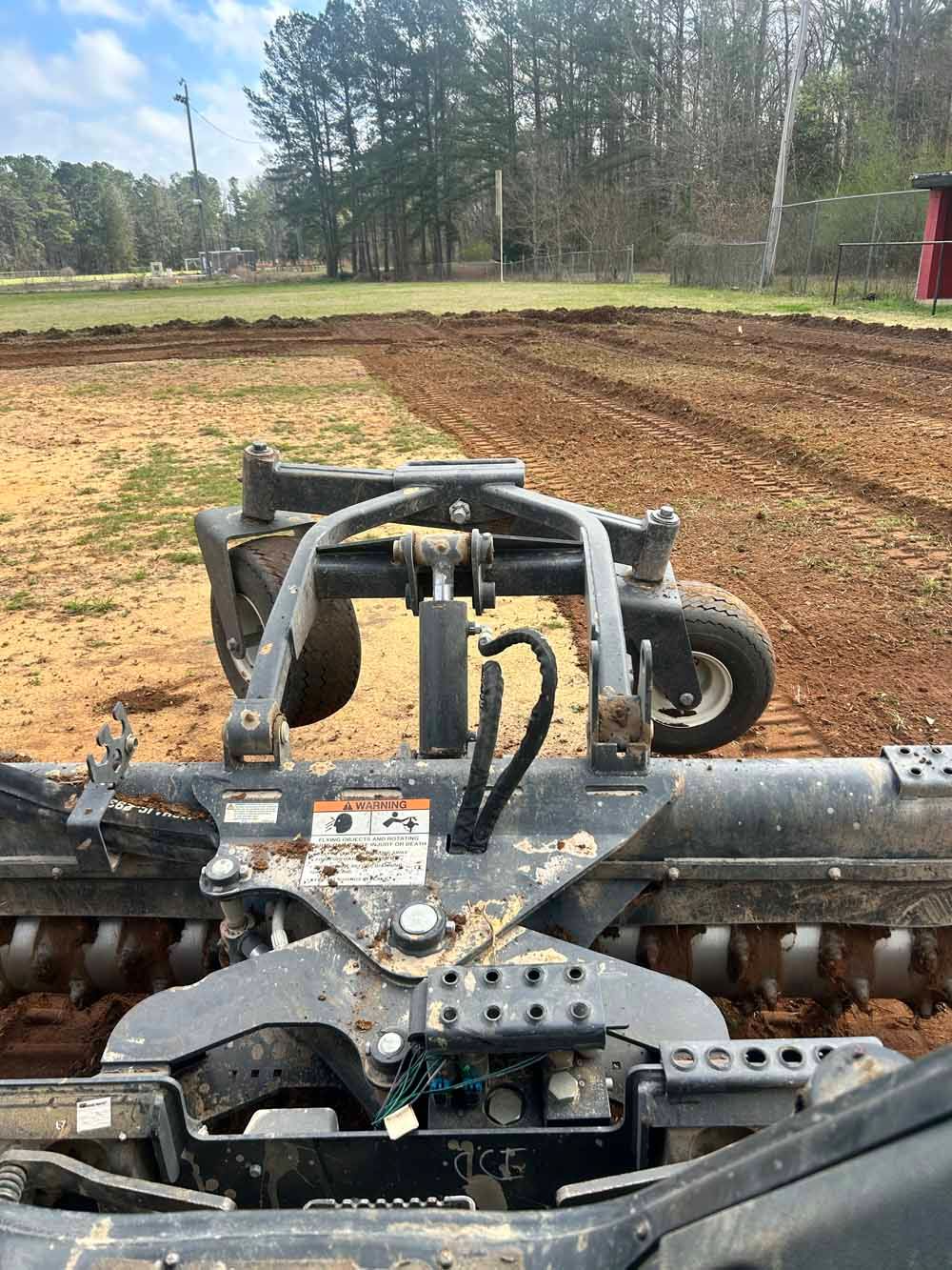 Agricultural tiller attachment working on a dirt field with trees in the background