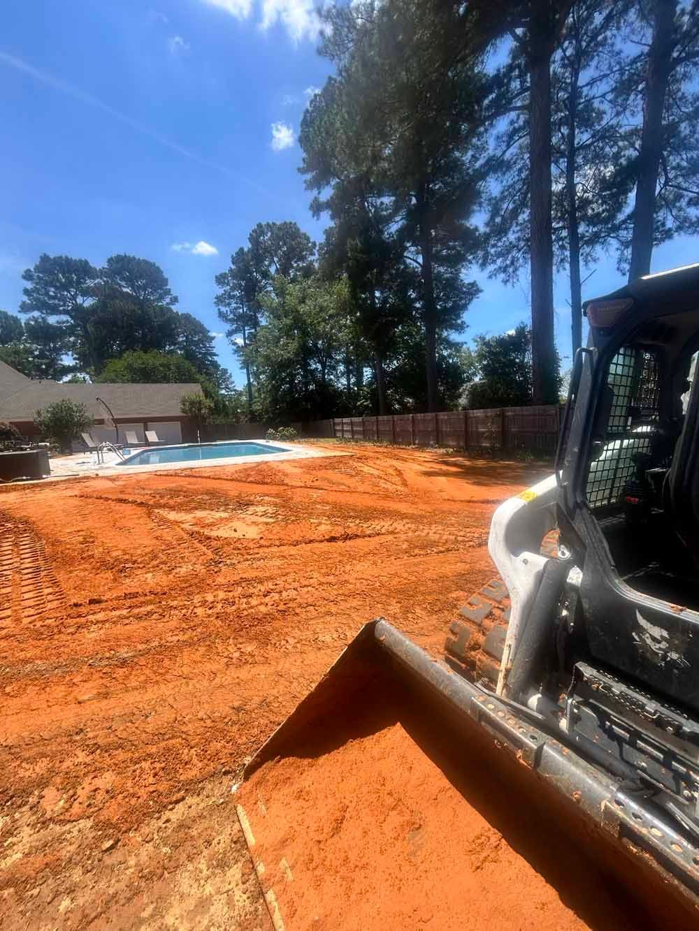 Bulldozer leveling red dirt on a sunny construction site with trees and a house nearby
