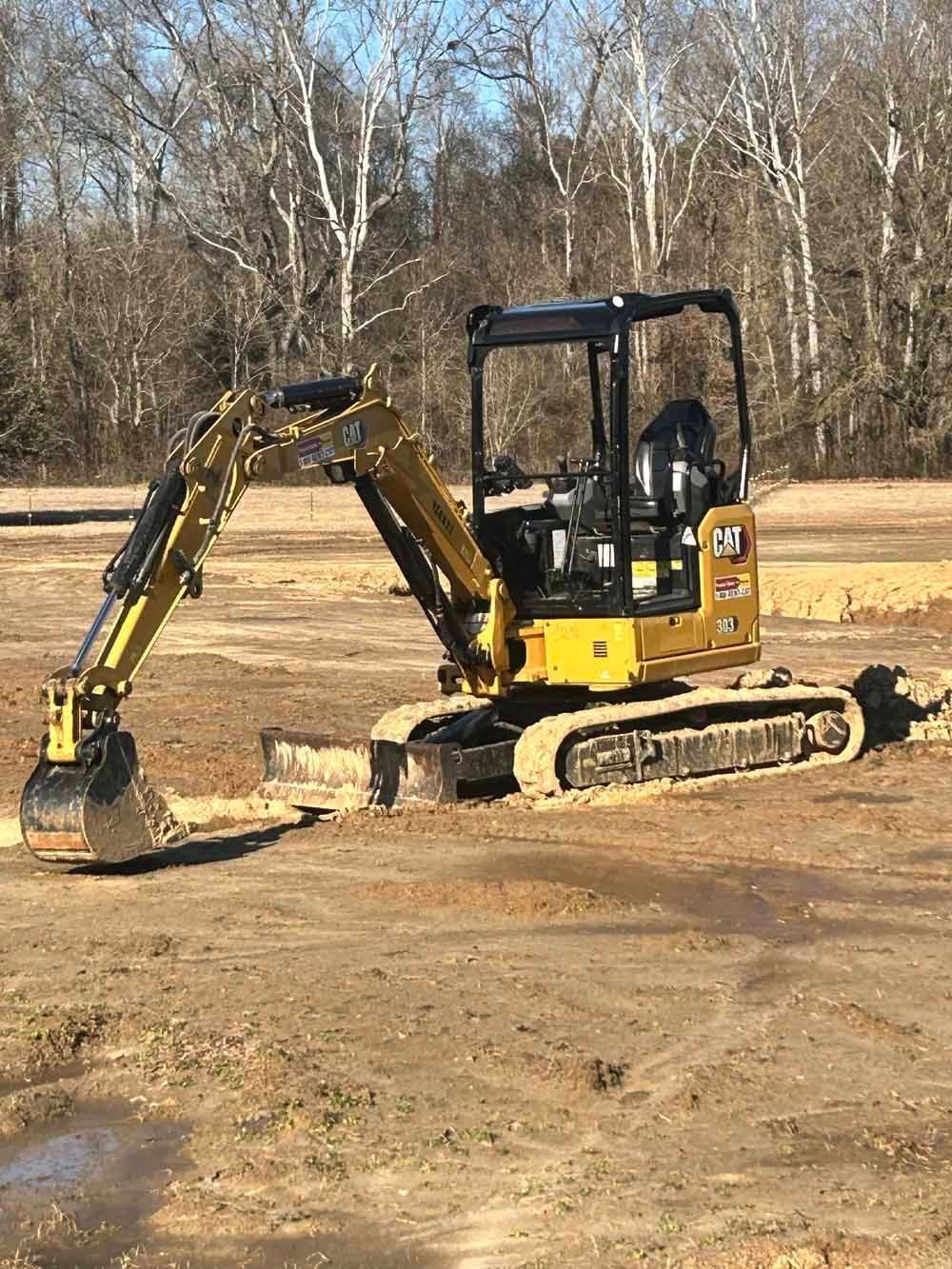 Yellow tracked excavator with raised arm in a dirt lot near leafless trees