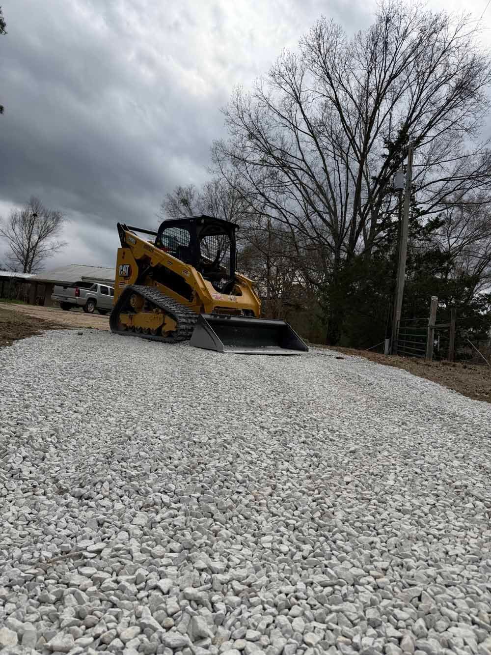 Yellow skid steer on a gravel mound under cloudy skies, with bare trees in the background