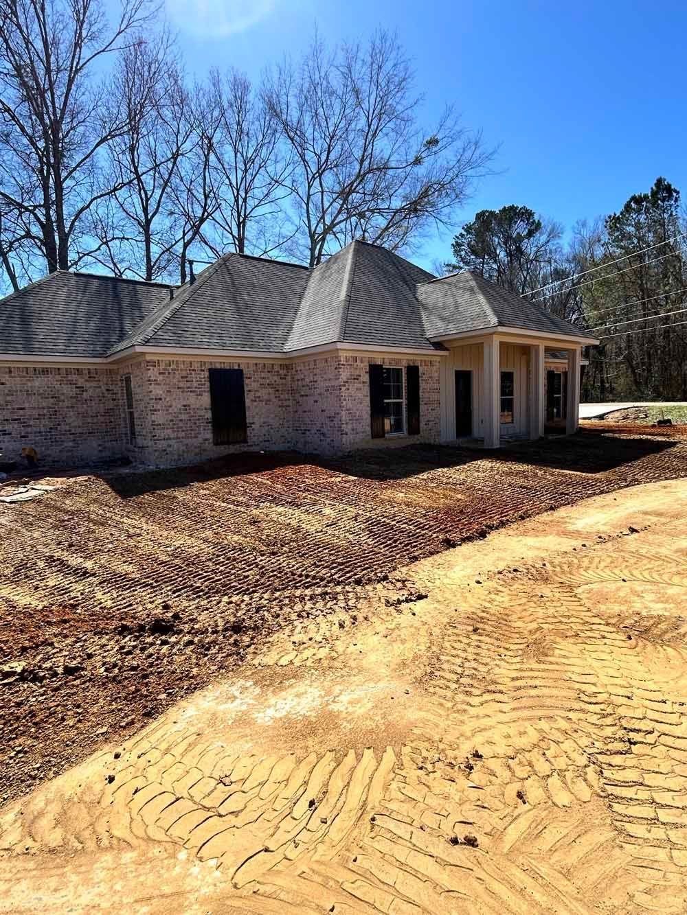 New brick house under construction with dark shingle roof and dirt driveway under blue sky