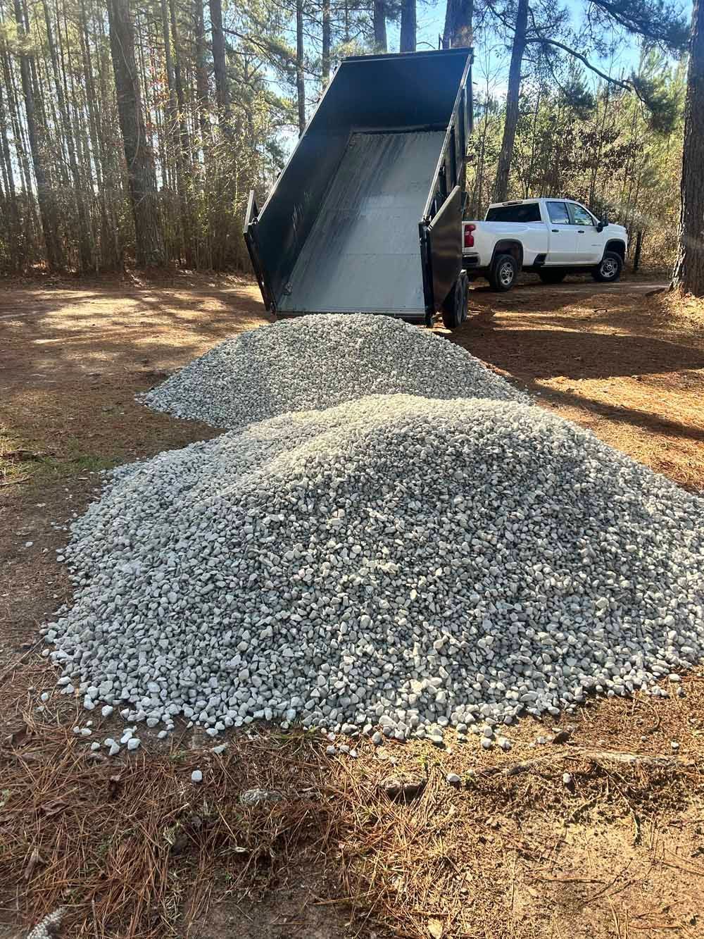 Dump truck unloading gravel into a large pile in a wooded area, with a white pickup nearby.