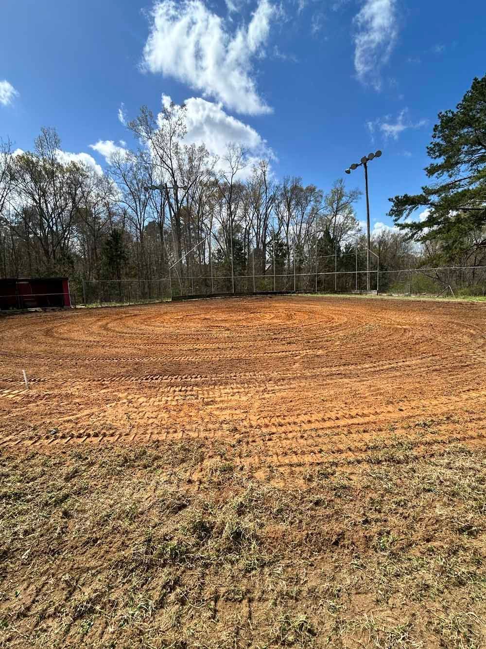 Freshly graded dirt field under a blue sky with clouds and trees in the background
