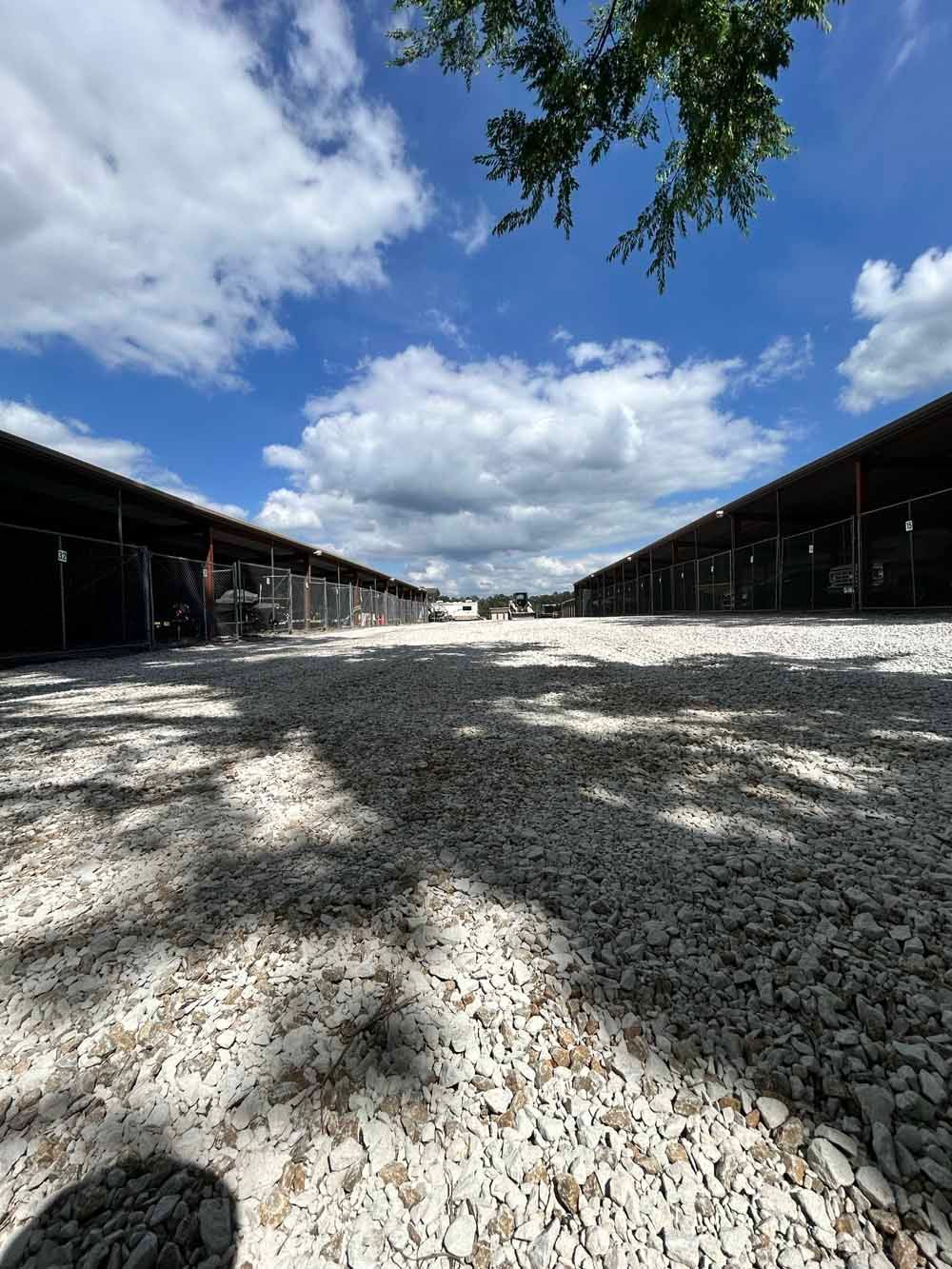 Open gravel courtyard between long dark-roofed barns under a bright blue sky with clouds.