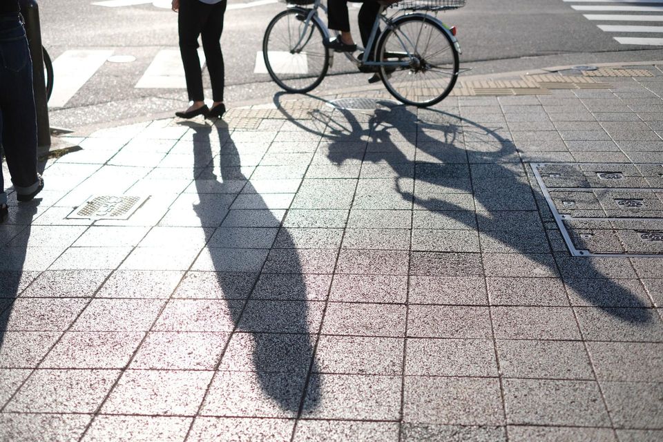 A person stands on a paved sidewalk next to another person riding a bicycle, with their long shadows cast on the ground.