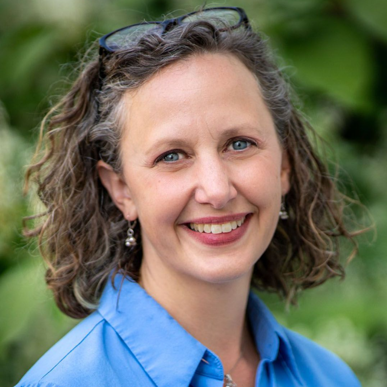 A smiling woman with wavy hair and glasses atop her head wears a blue collared shirt against a blurred greenery background.