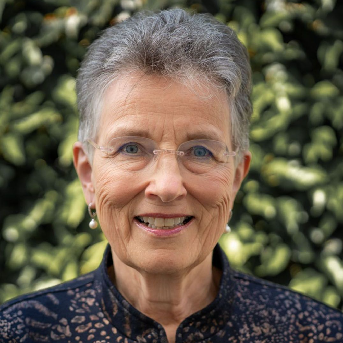 A person with short, gray hair and glasses smiles while wearing a dark, patterned top in front of a leafy green background.