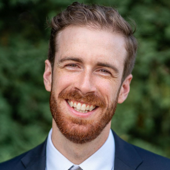 A person with a beard and light brown hair smiling at the camera, wearing a suit and tie against a blurred green backdrop.