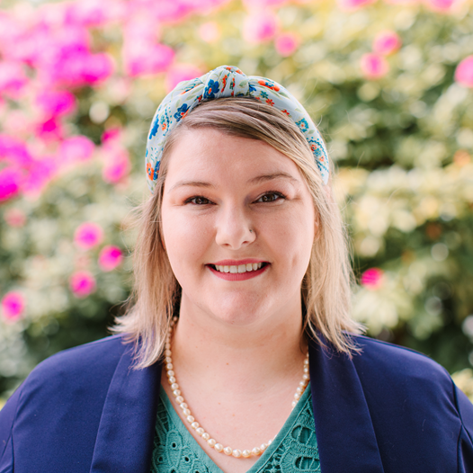 A person with medium light brown hair wearing a blue floral headband, a pearl necklace, teal lace shirt and blue blazer against a leafy backdrop.