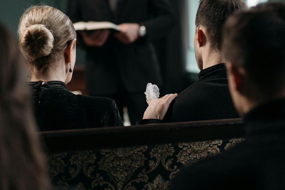 Attendees in dark clothing sit in church pews during a service, with one person resting a hand on another's shoulder.