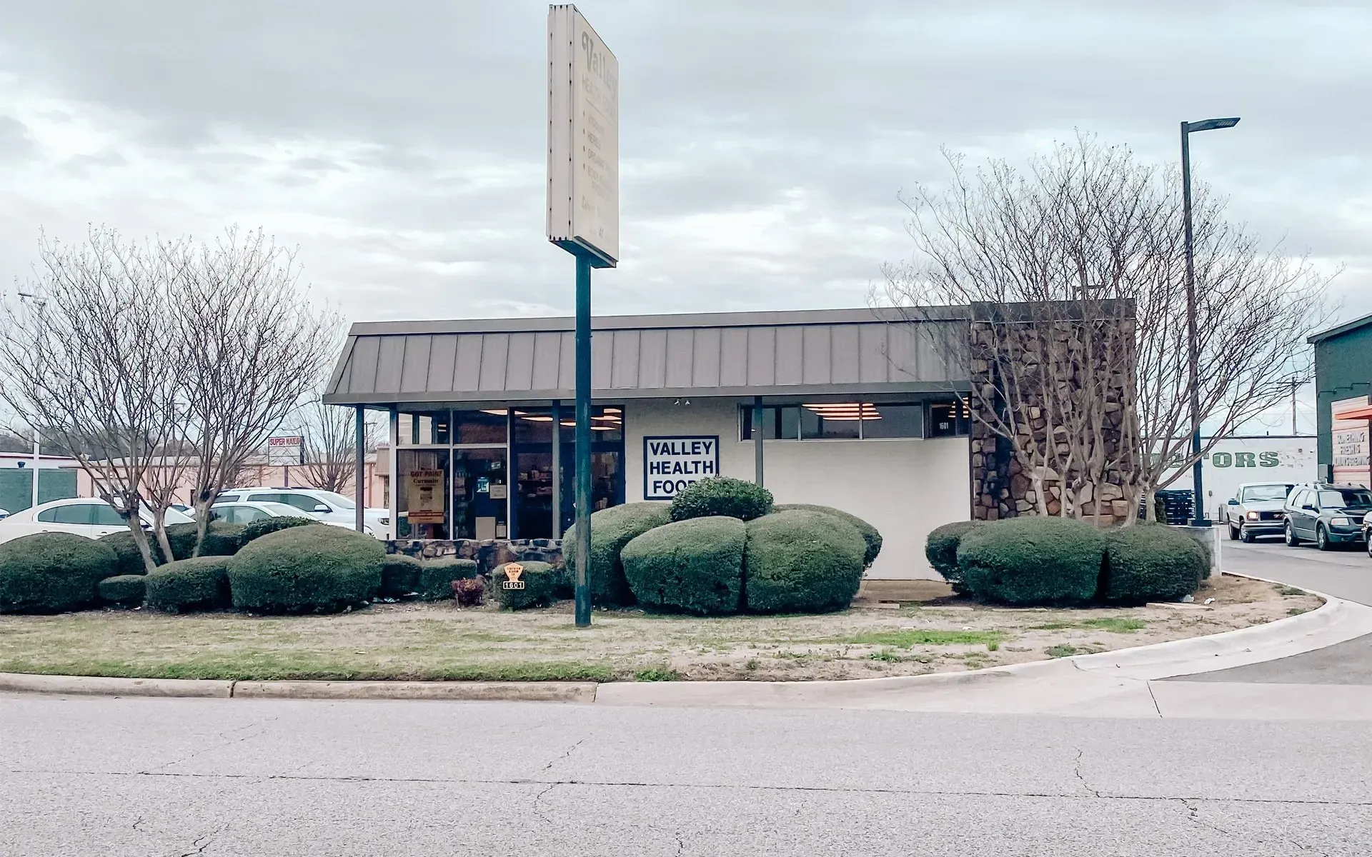 Small, light-colored building with a dark roof and a sign, surrounded by shrubs and bare trees on a cloudy day. Small, light-colored building with a dark roof and a sign, surrounded by shrubs and bare trees on a cloudy day.