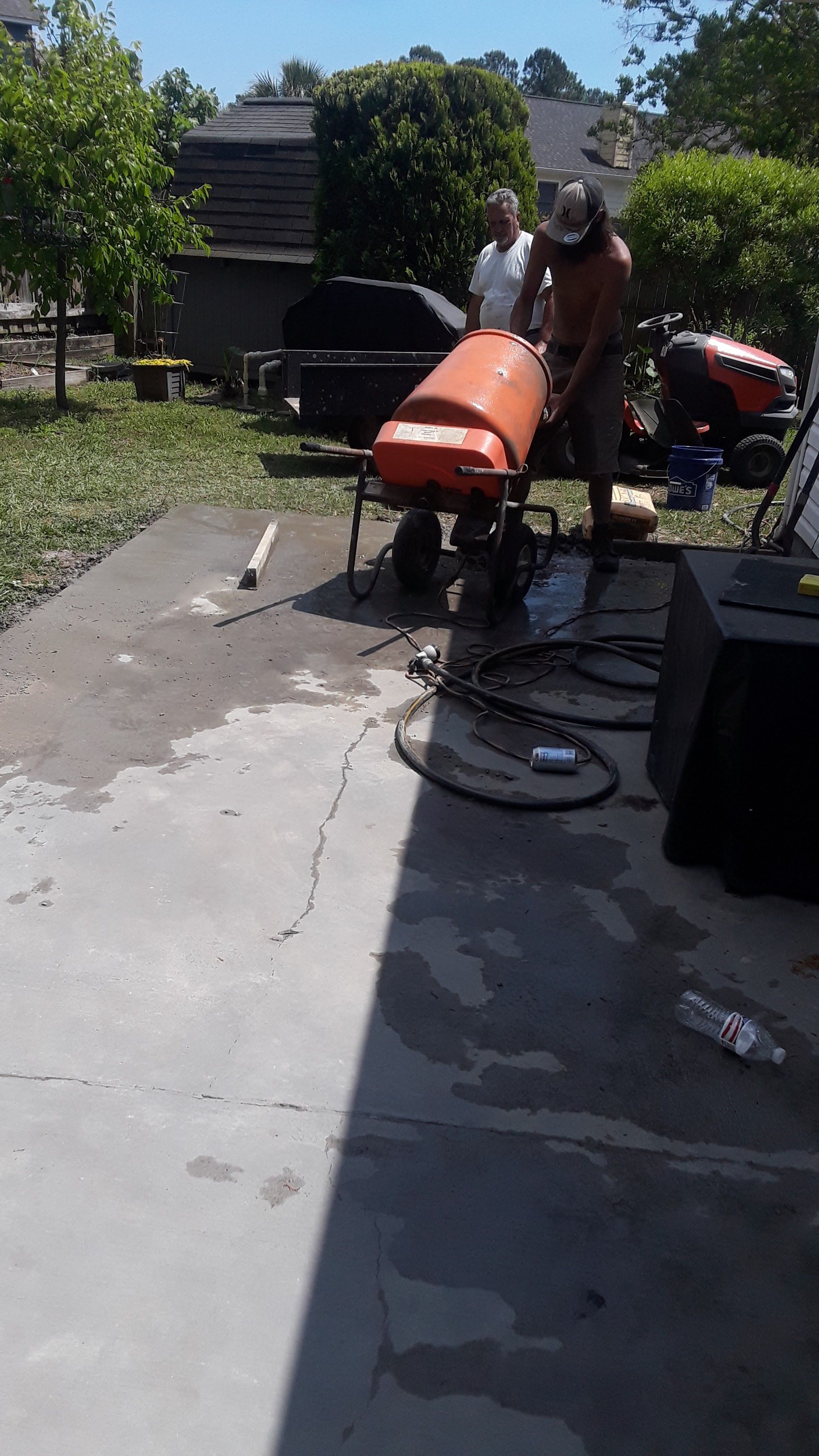 Two people stand on a concrete patio, one operating a pressurized orange tank on wheels while water sprays on the ground.