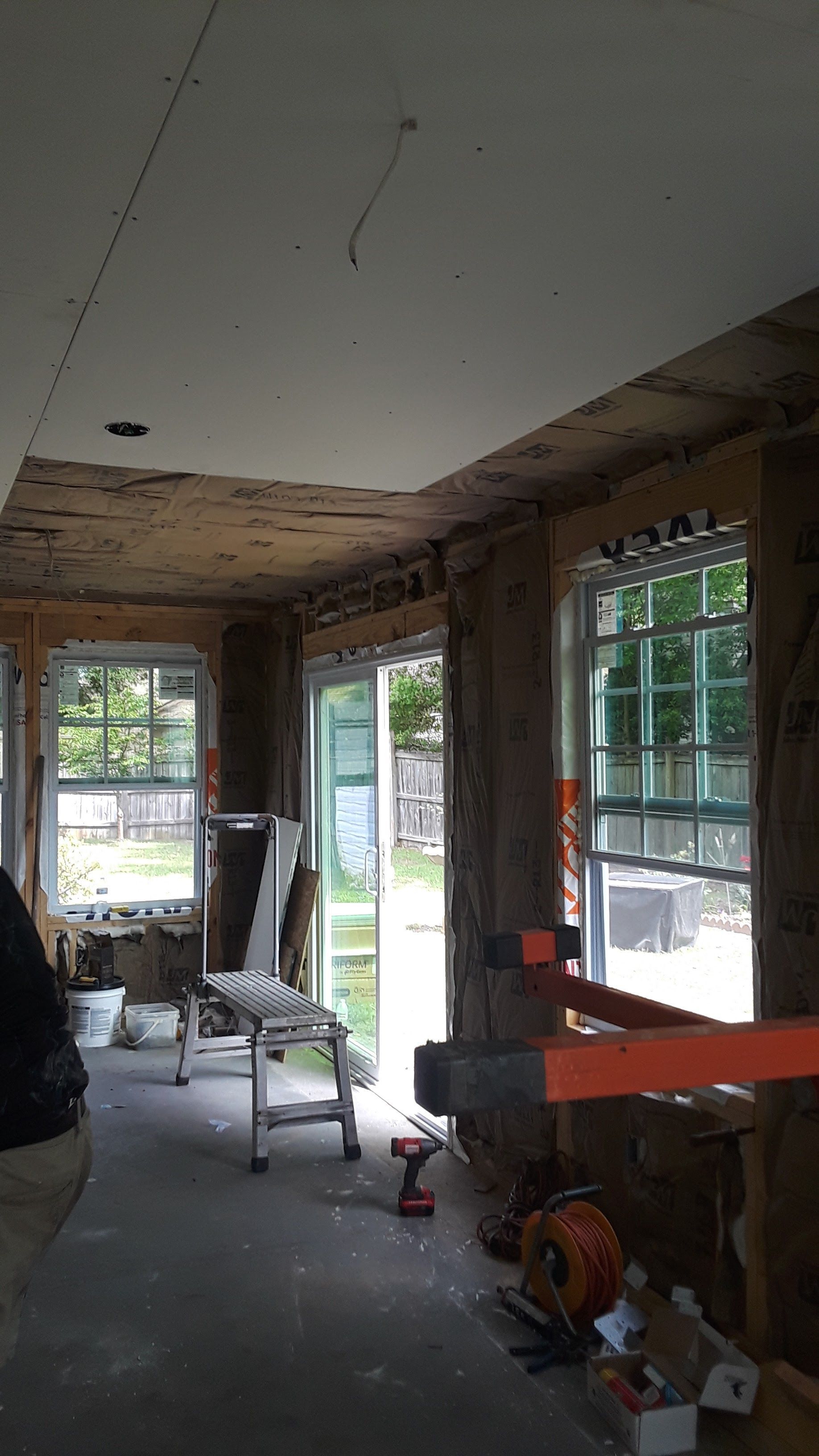 Interior view of a residential room under construction, featuring exposed wooden studs, drywall, and large windows.