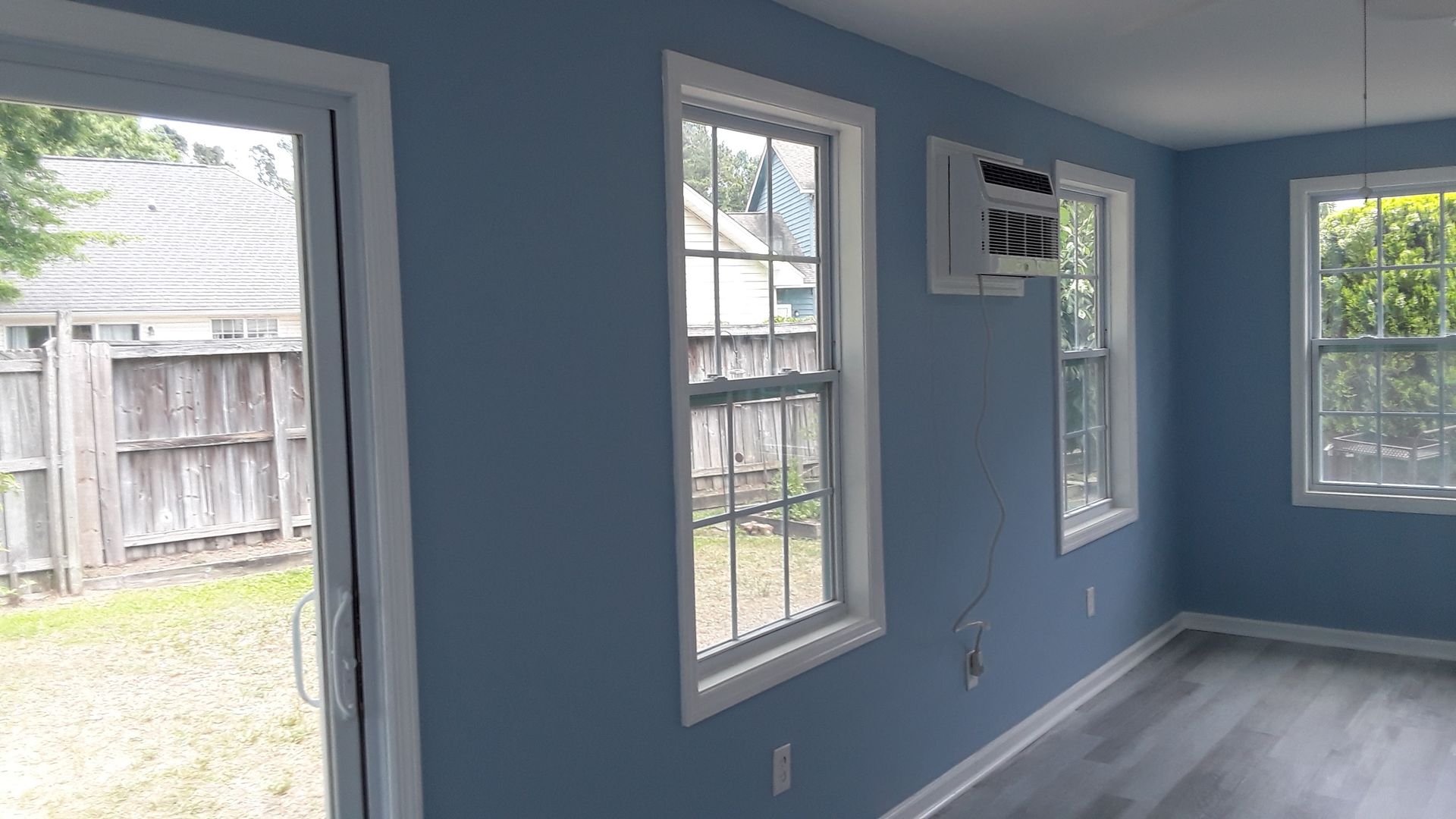 Interior view of a room with light blue walls, gray flooring, a sliding glass door, and two windows with an AC unit.