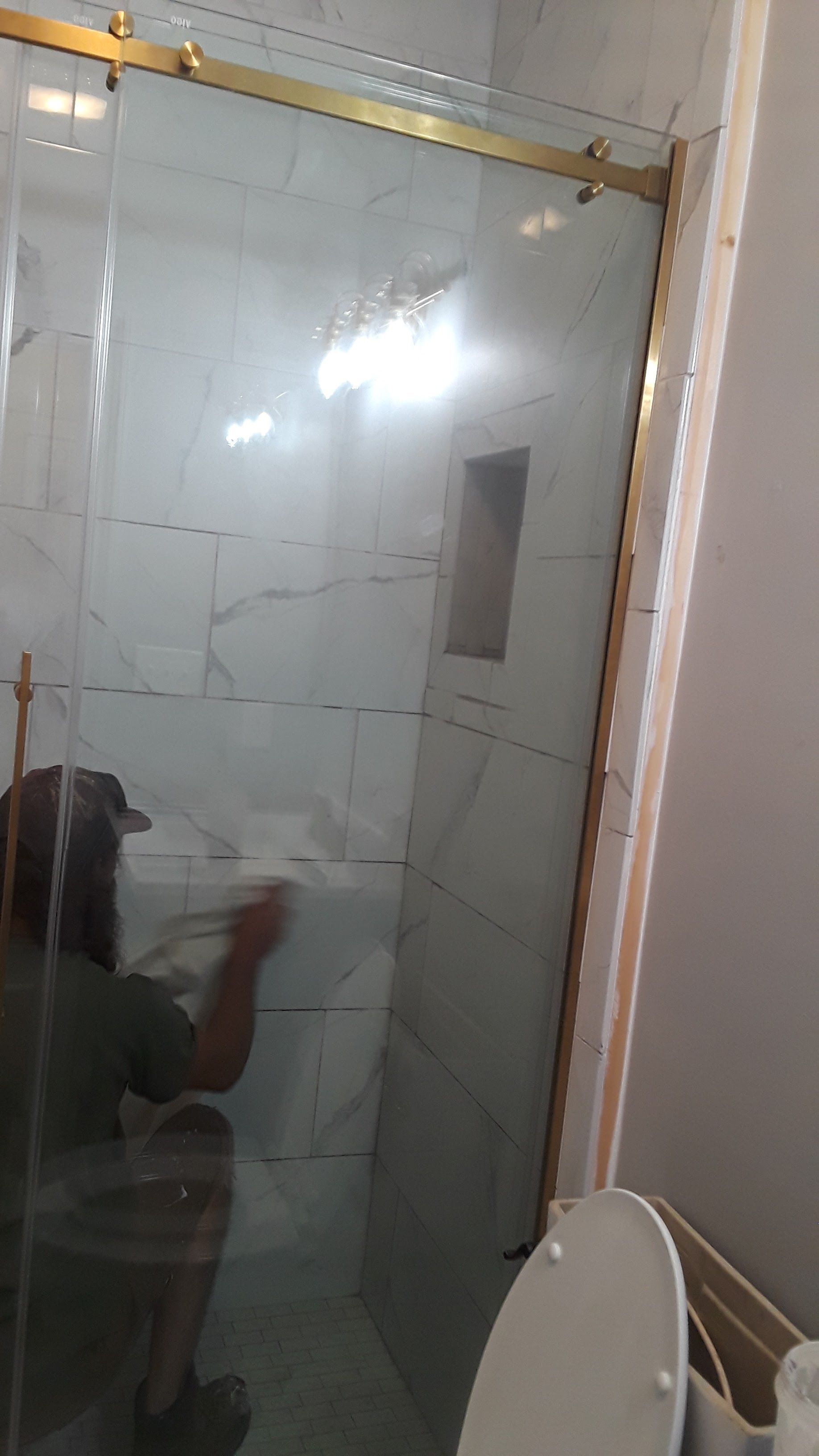 A person wiping down a glass shower door with gold hardware in a bathroom with white marble tile.