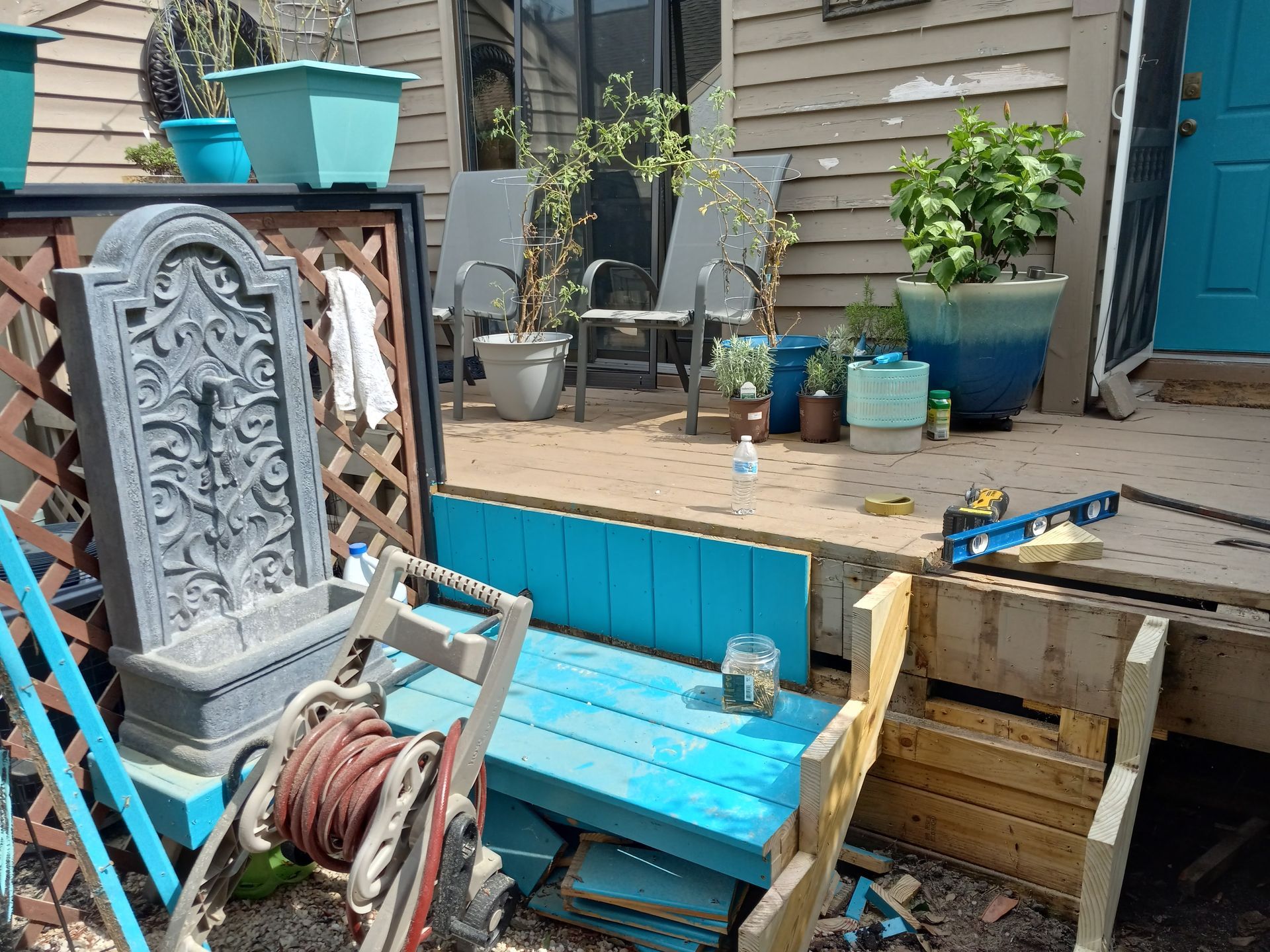 A grey wall fountain stands near a blue DIY bench and a hose reel on a backyard patio with potted plants and wooden steps.