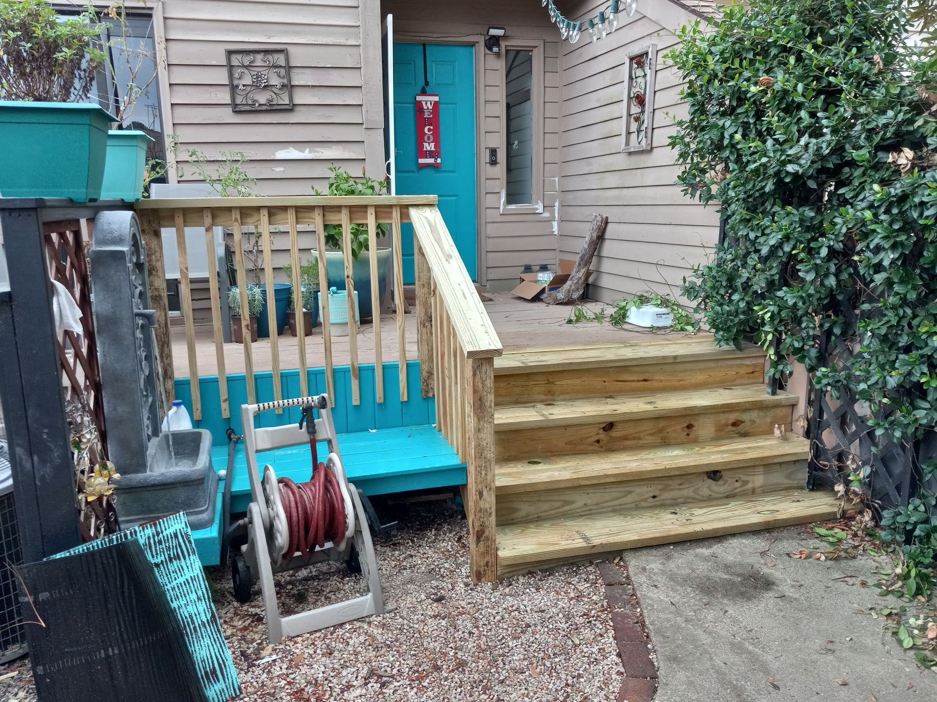 New wooden porch steps lead up to a bright turquoise door on a tan house with a small blue bench on the side.