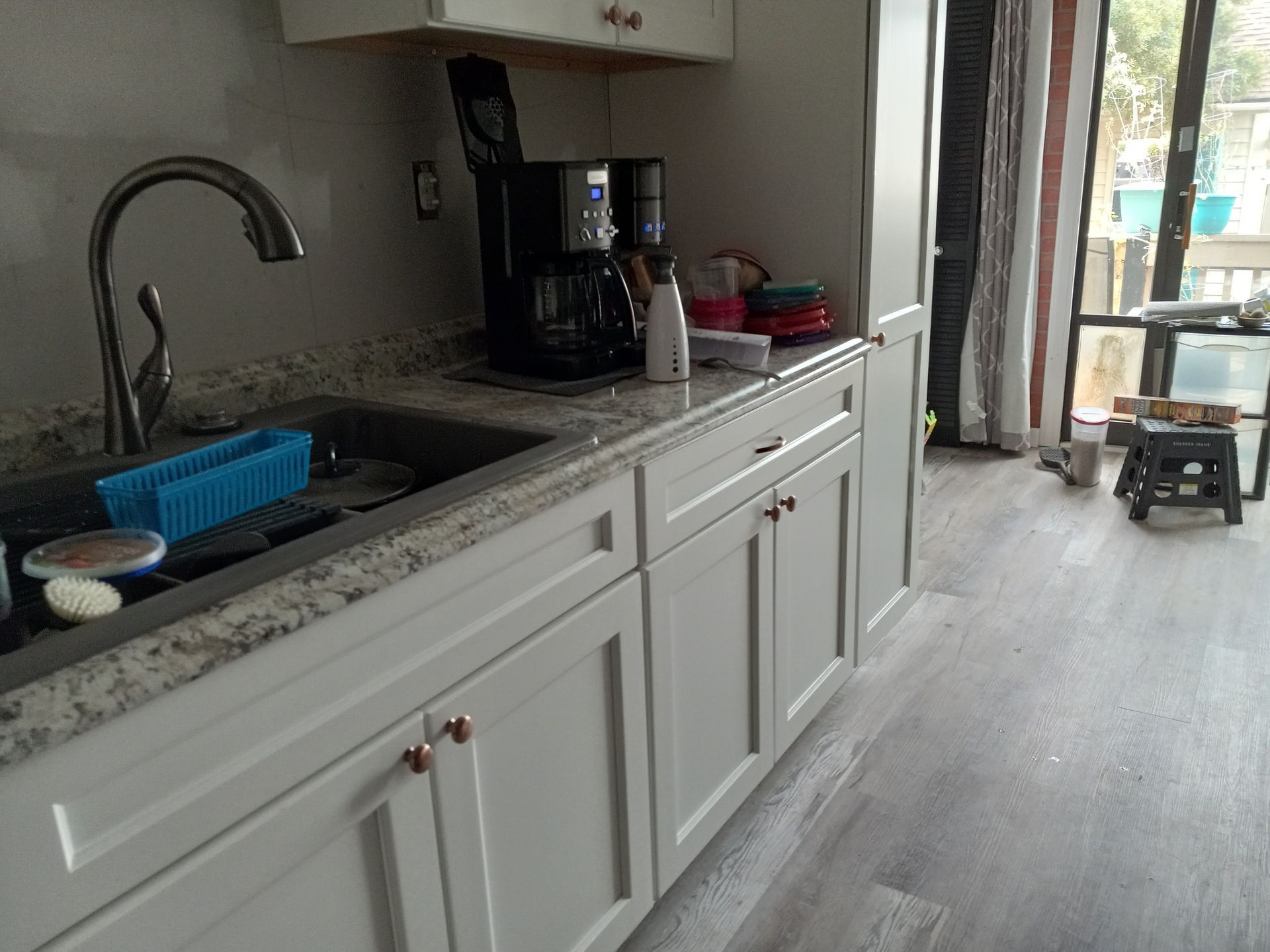 A kitchen counter with a sink, coffee maker, and white cabinets, with a doorway leading to an outdoor space.