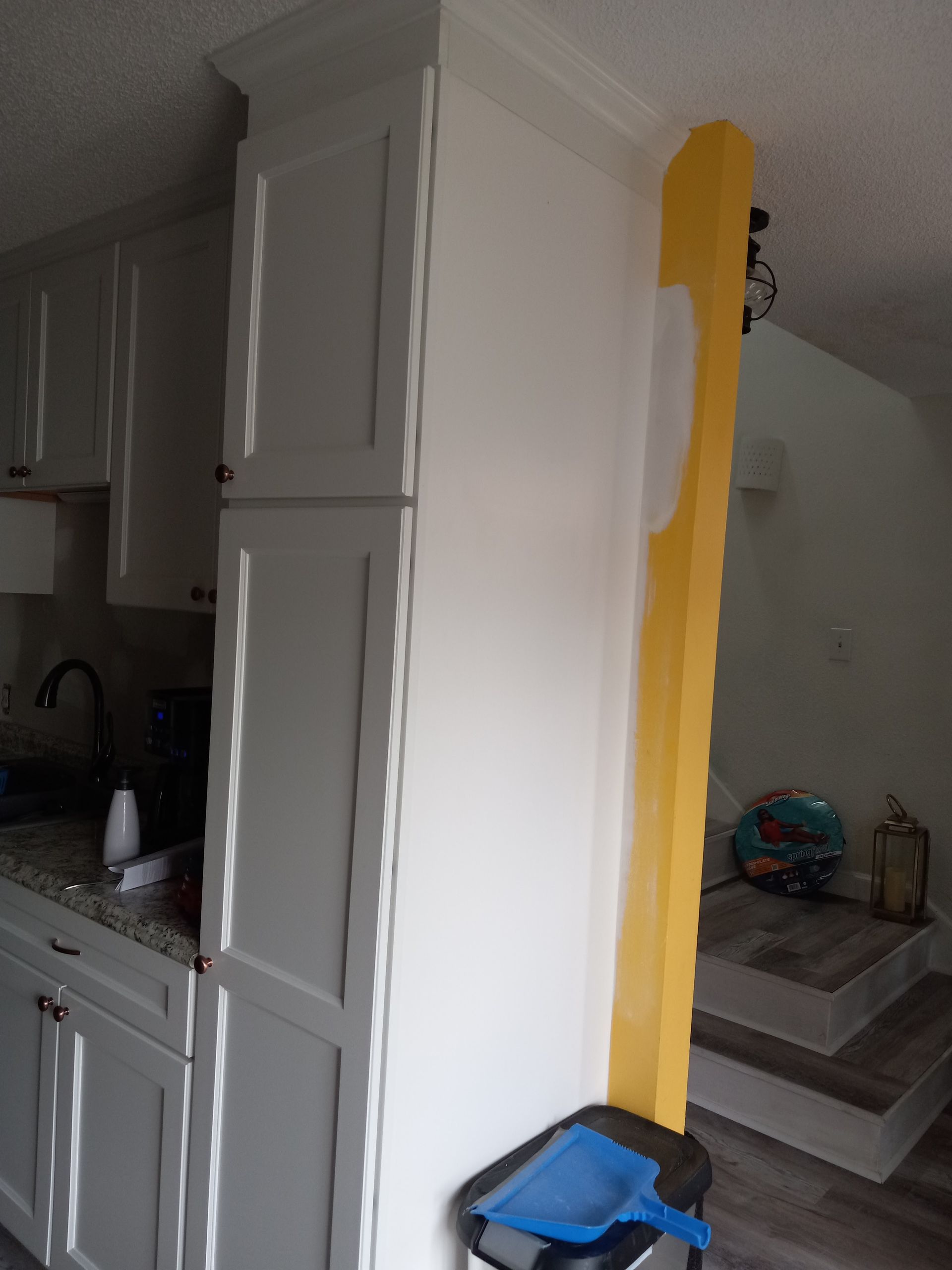 A white kitchen cabinet stands beside a yellow wall column that has been partially painted white.