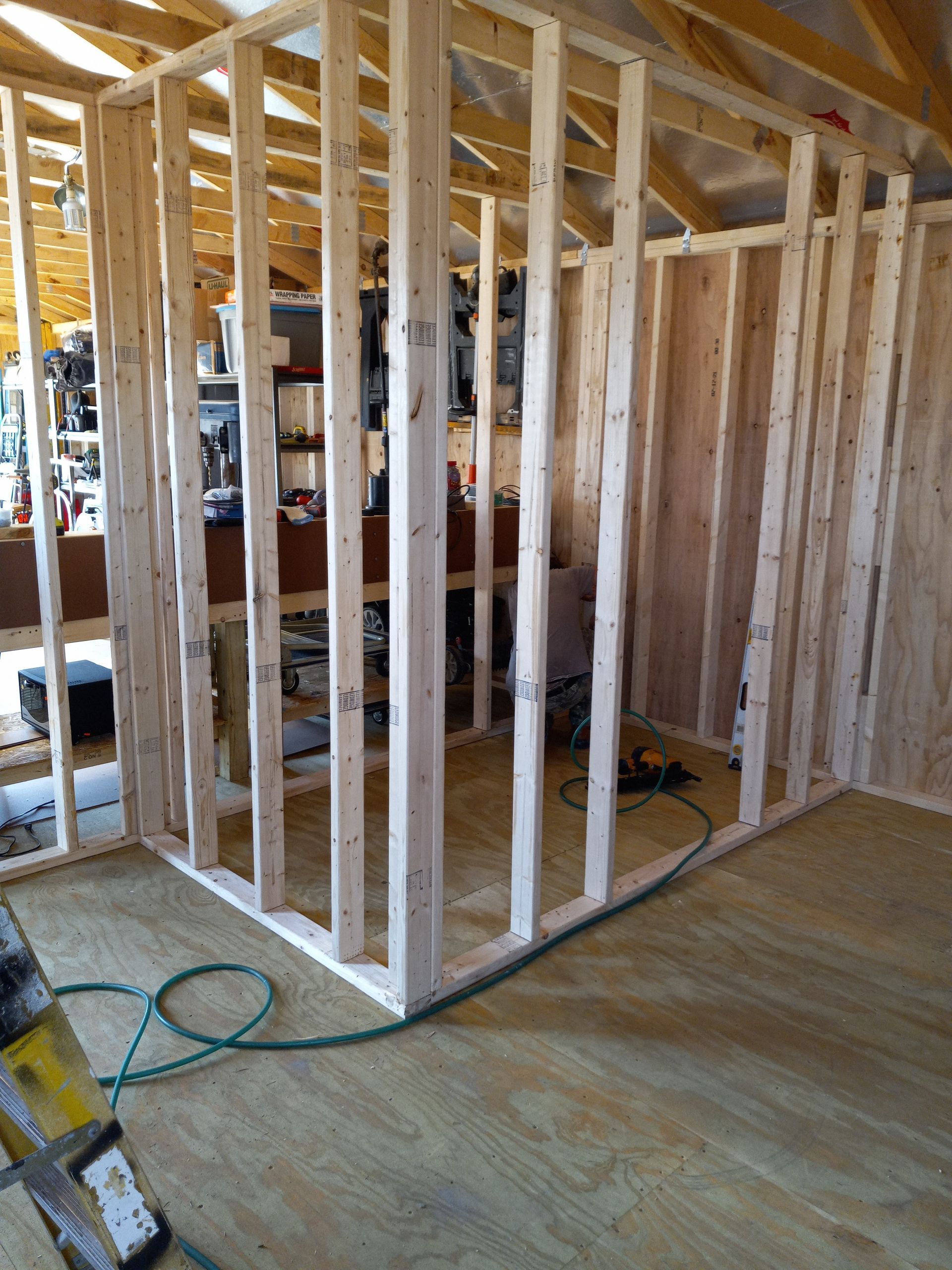 Wooden wall framing for a room under construction inside a shed, showing vertical studs on a plywood floor.