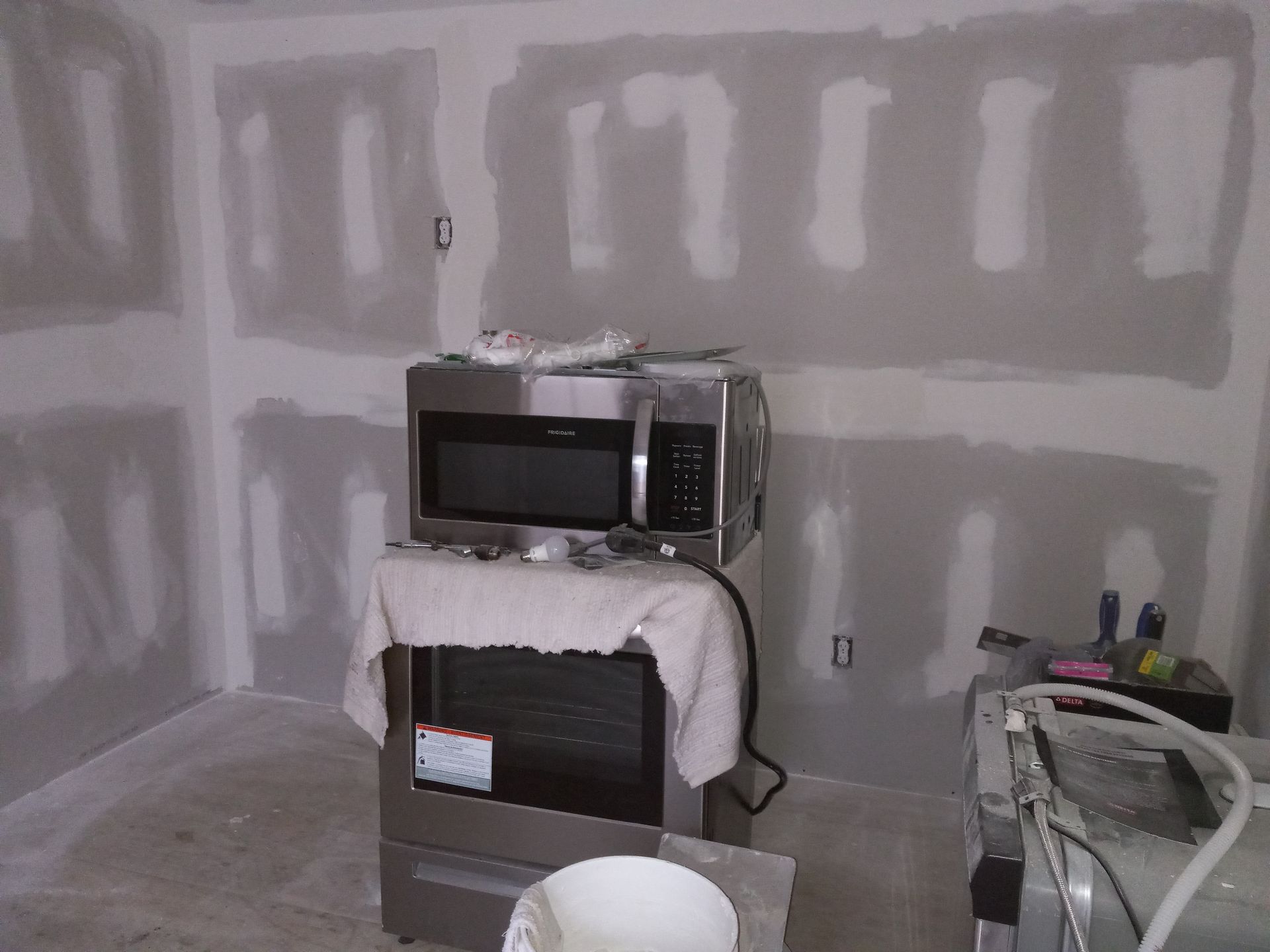 A stainless-steel microwave and oven unit sits in a room with unfinished drywall walls and exposed electrical outlets.