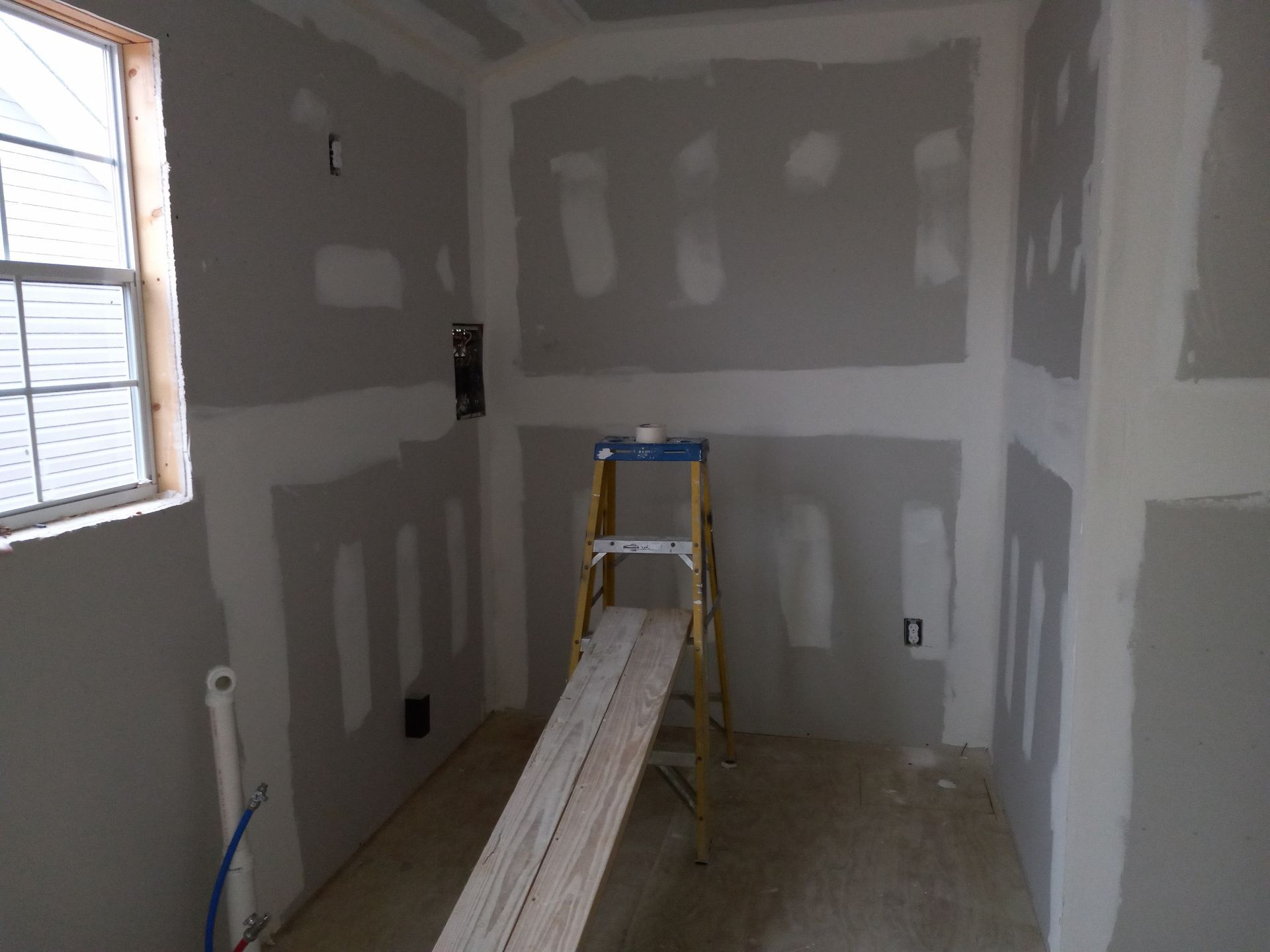 An interior room under construction with bare drywall, a window, and a ladder with a plank positioned in the center.