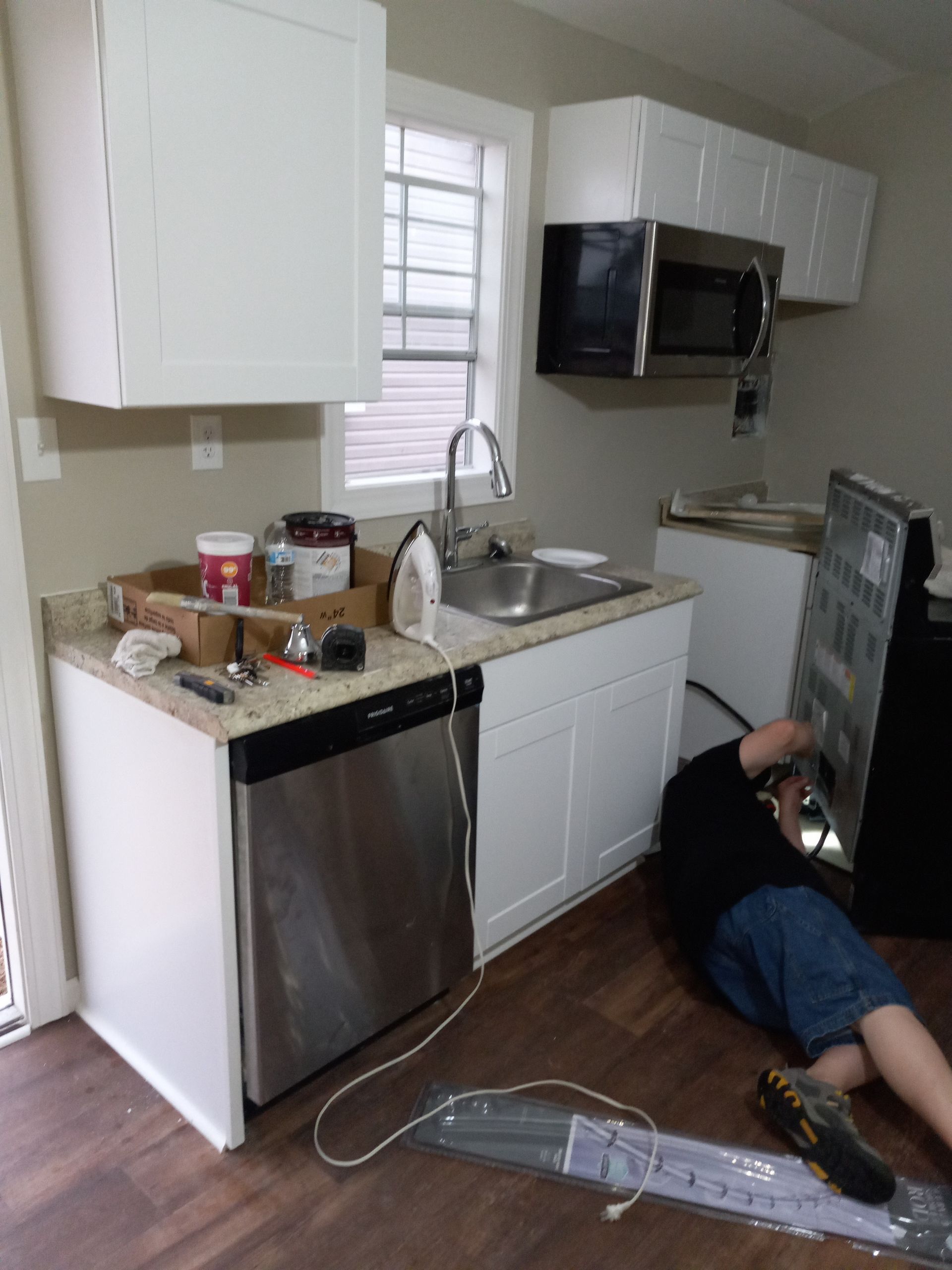 A person works on the back of a black refrigerator in a kitchen with white cabinets, a sink, and a stainless dishwasher.