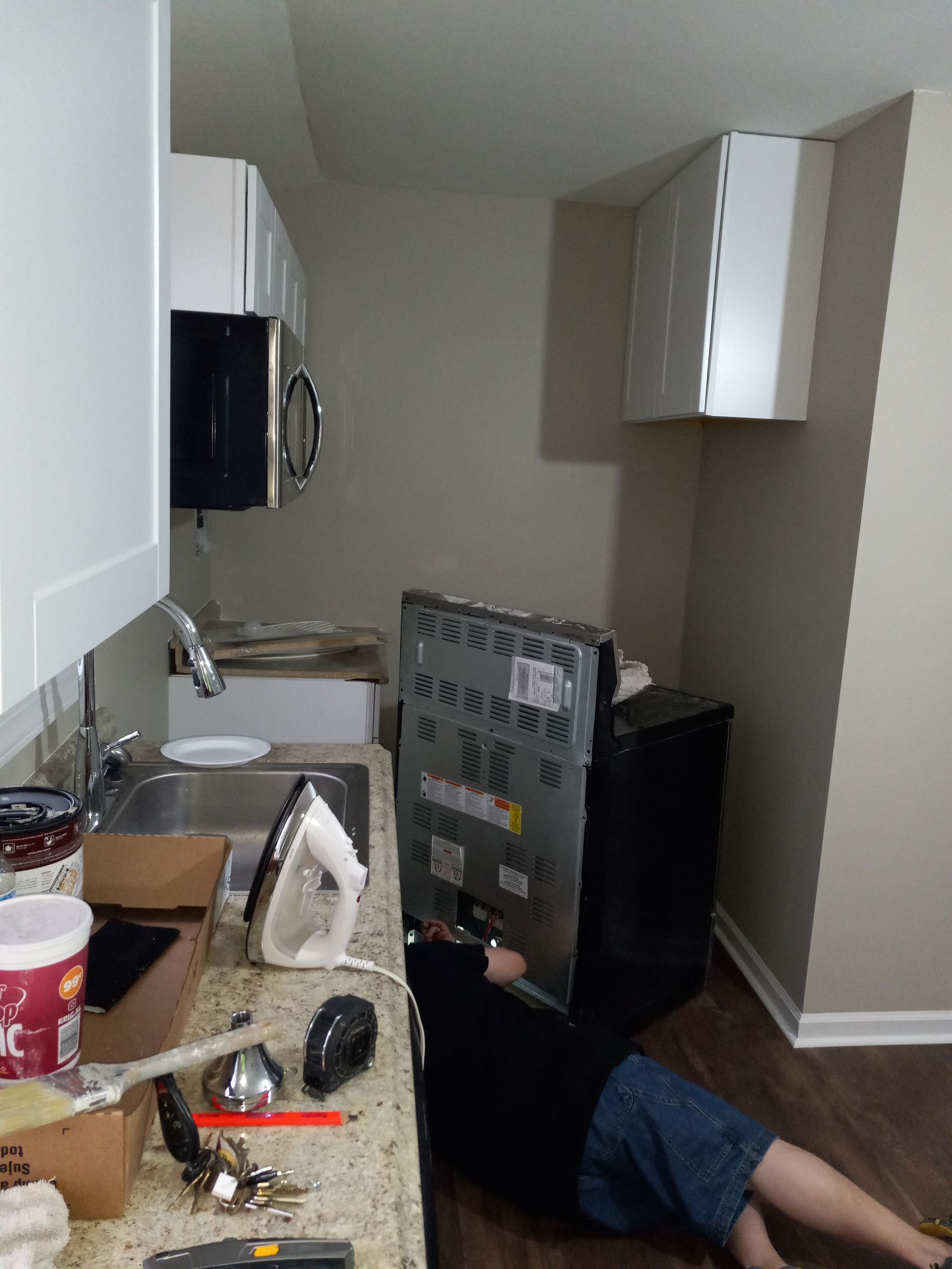 A person lying on a kitchen floor, reaching behind a black range to install or repair its electrical connection.