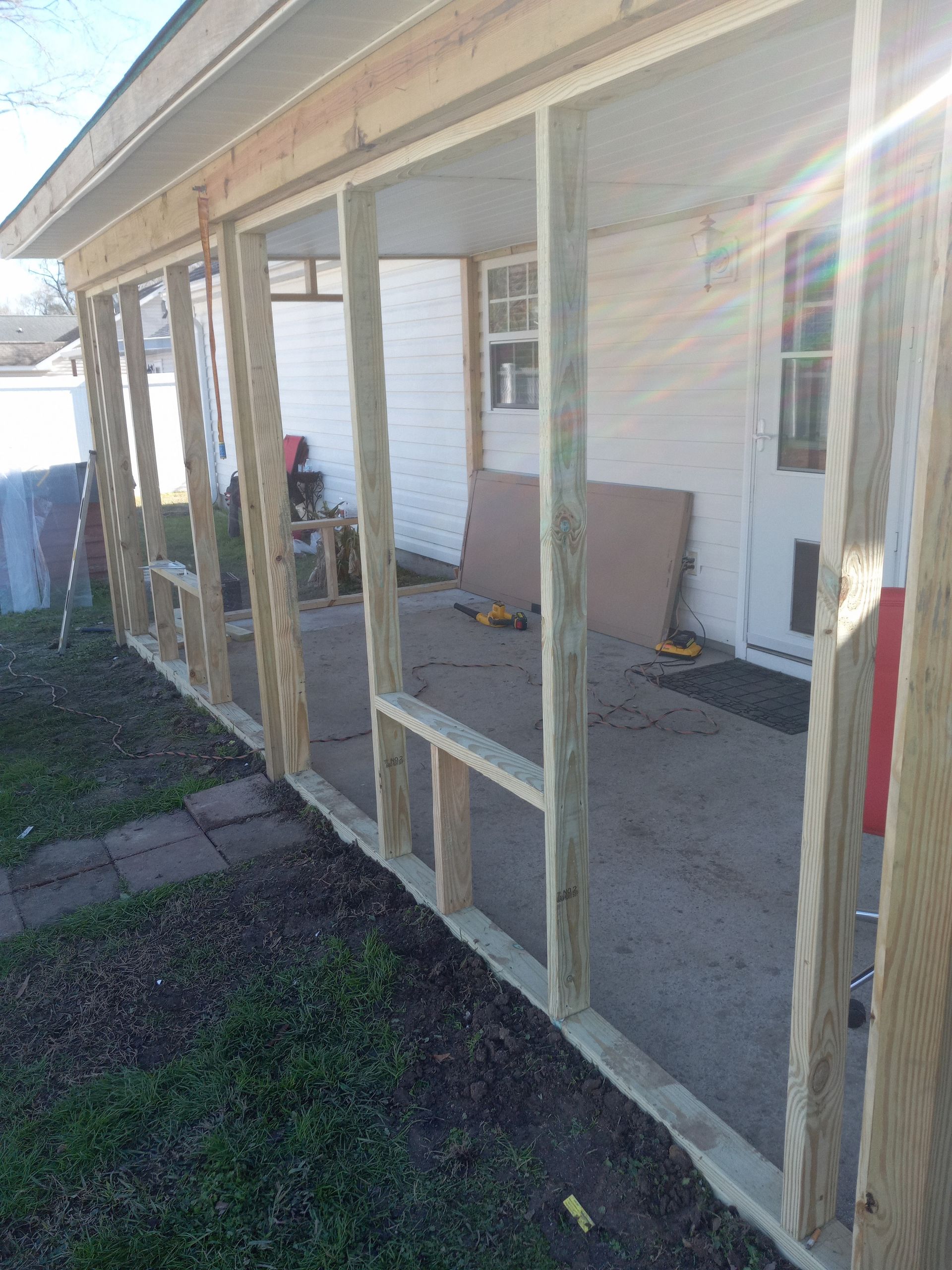 A wooden frame for a new porch wall stands on a concrete patio in front of a white house.
