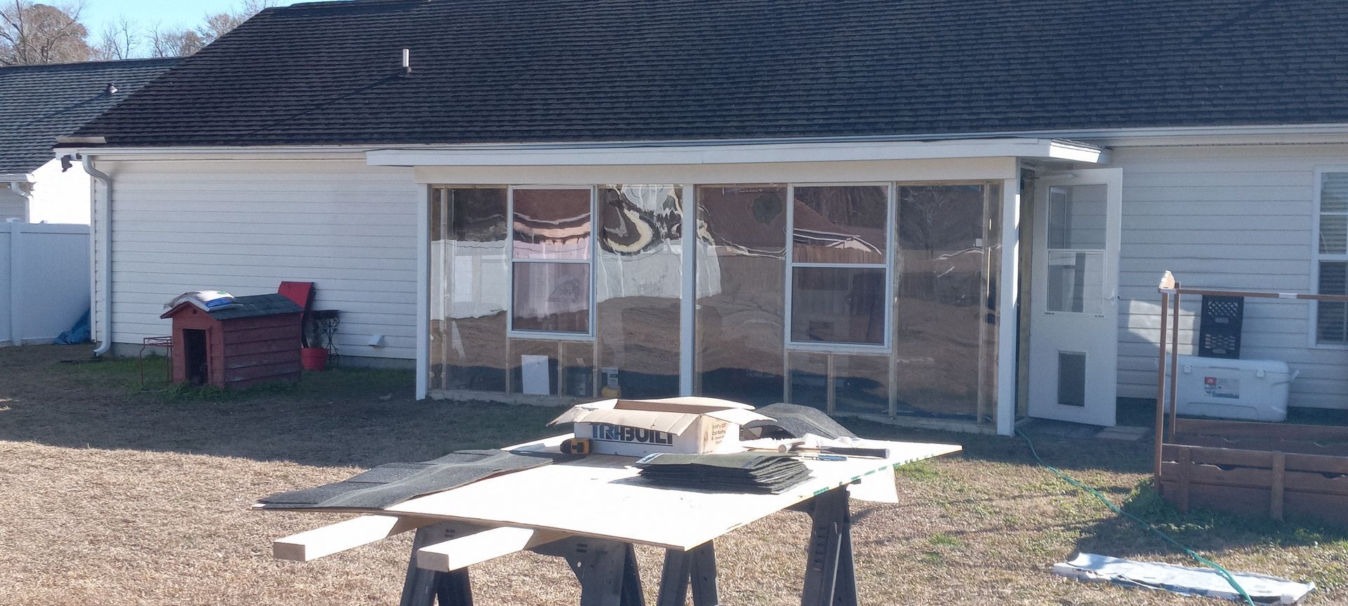 A house exterior with a sunroom under construction, featuring white siding, a red doghouse, and a workbench in the yard.