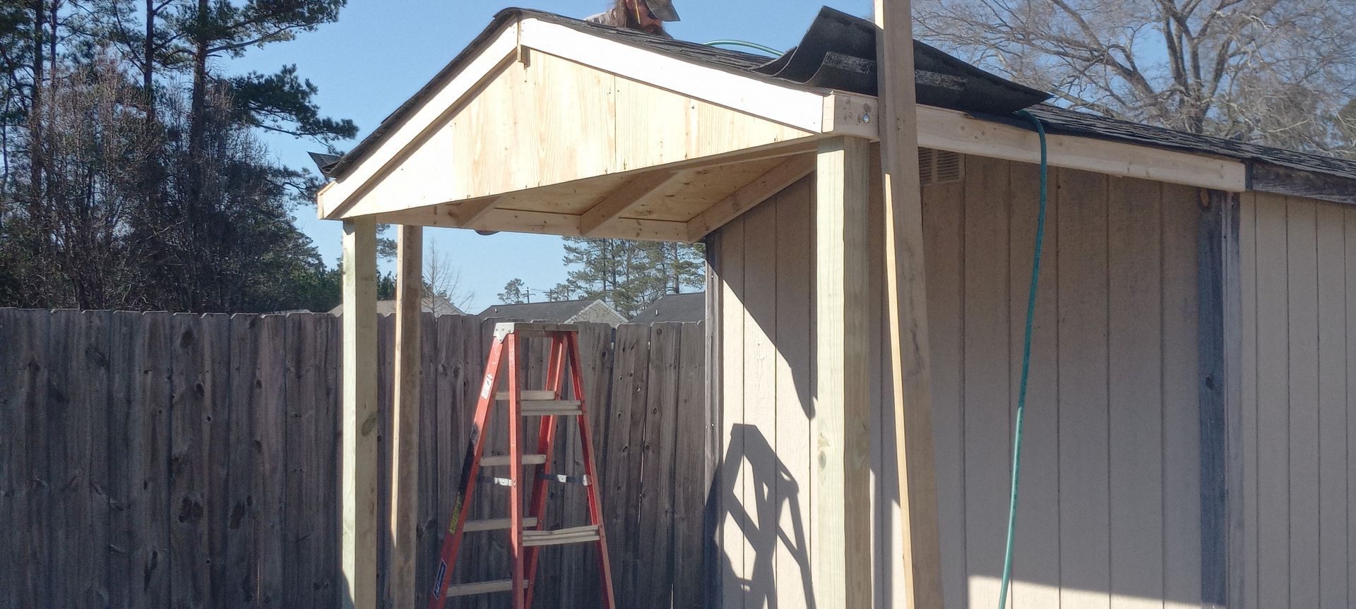 A partially constructed wooden porch roof attached to the side of a beige shed, with a red ladder standing underneath.