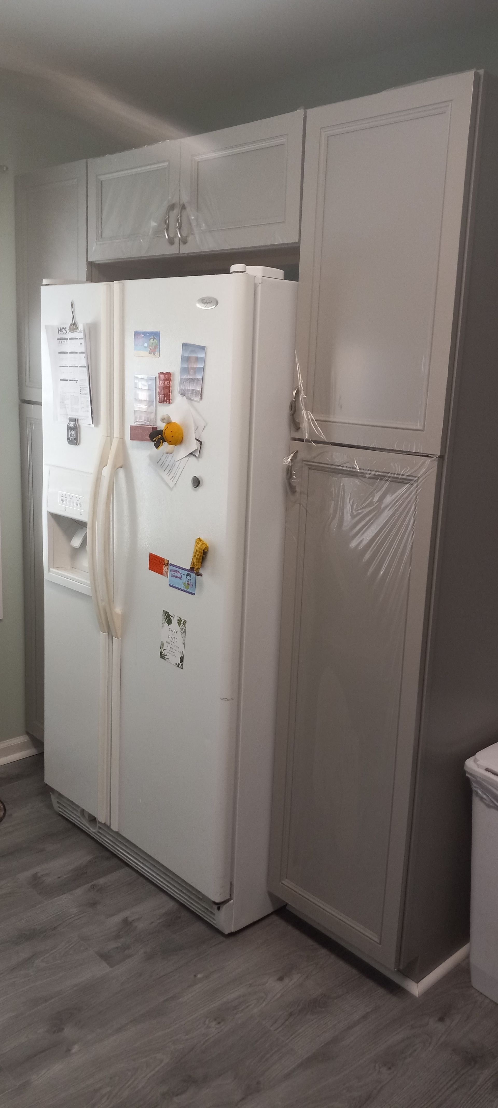 A white side-by-side refrigerator sits in a kitchen cabinet enclosure next to a tall, narrow grey pantry cabinet.