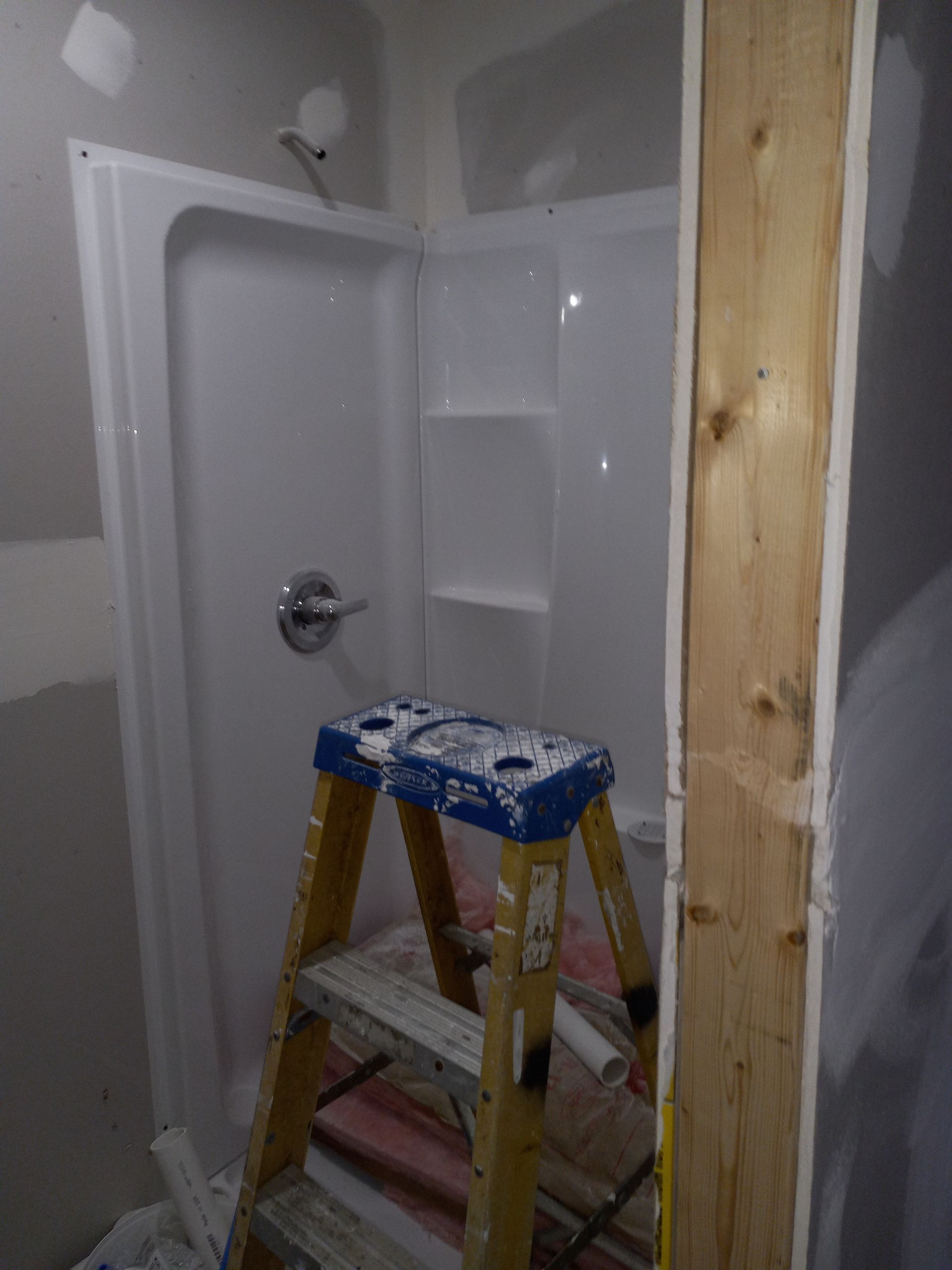 A yellow step ladder sits in a bathroom with an installed white shower surround and unfinished drywall.