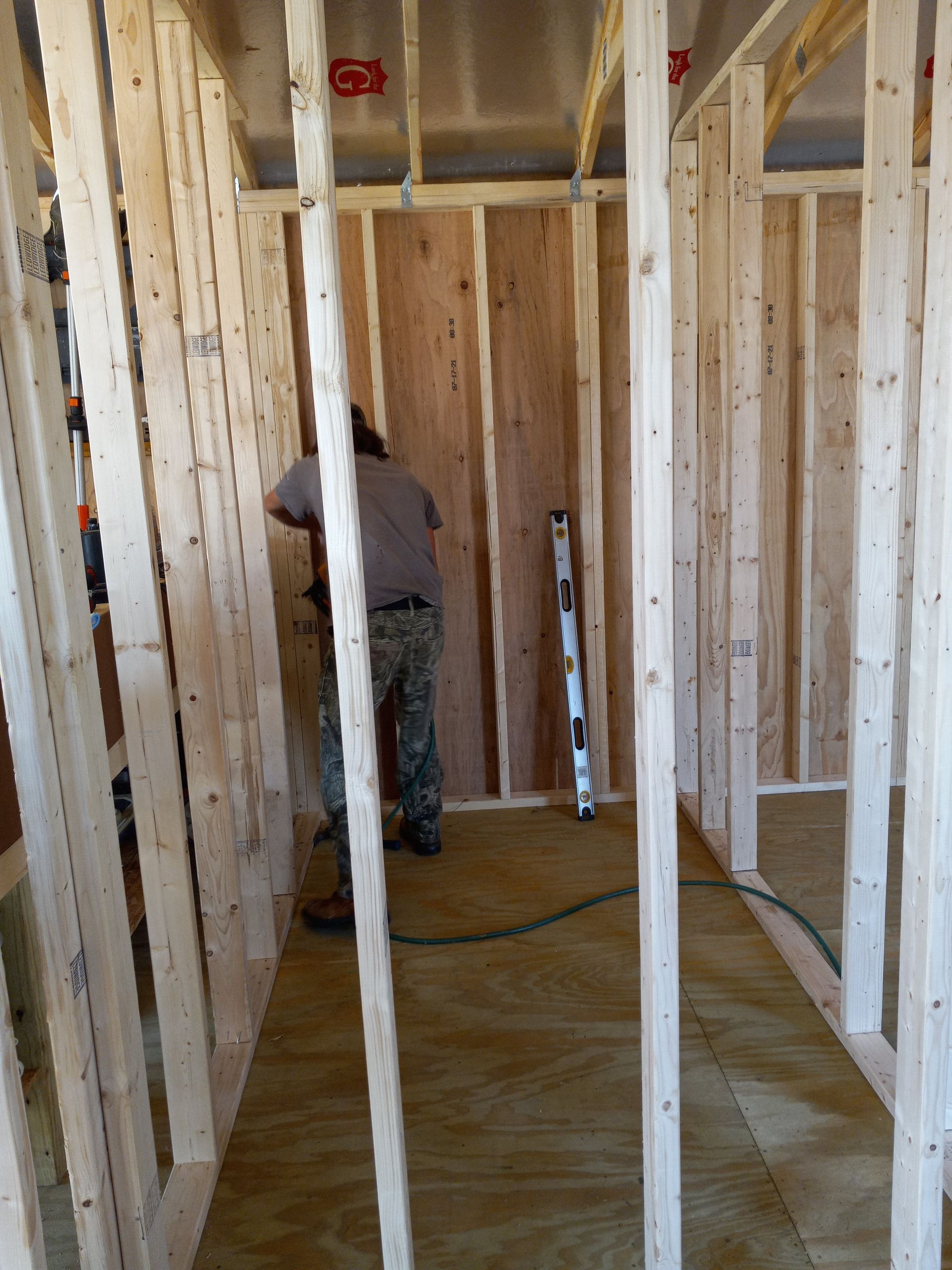 A person in work clothing stands inside a wooden frame construction site, working on the interior wall.
