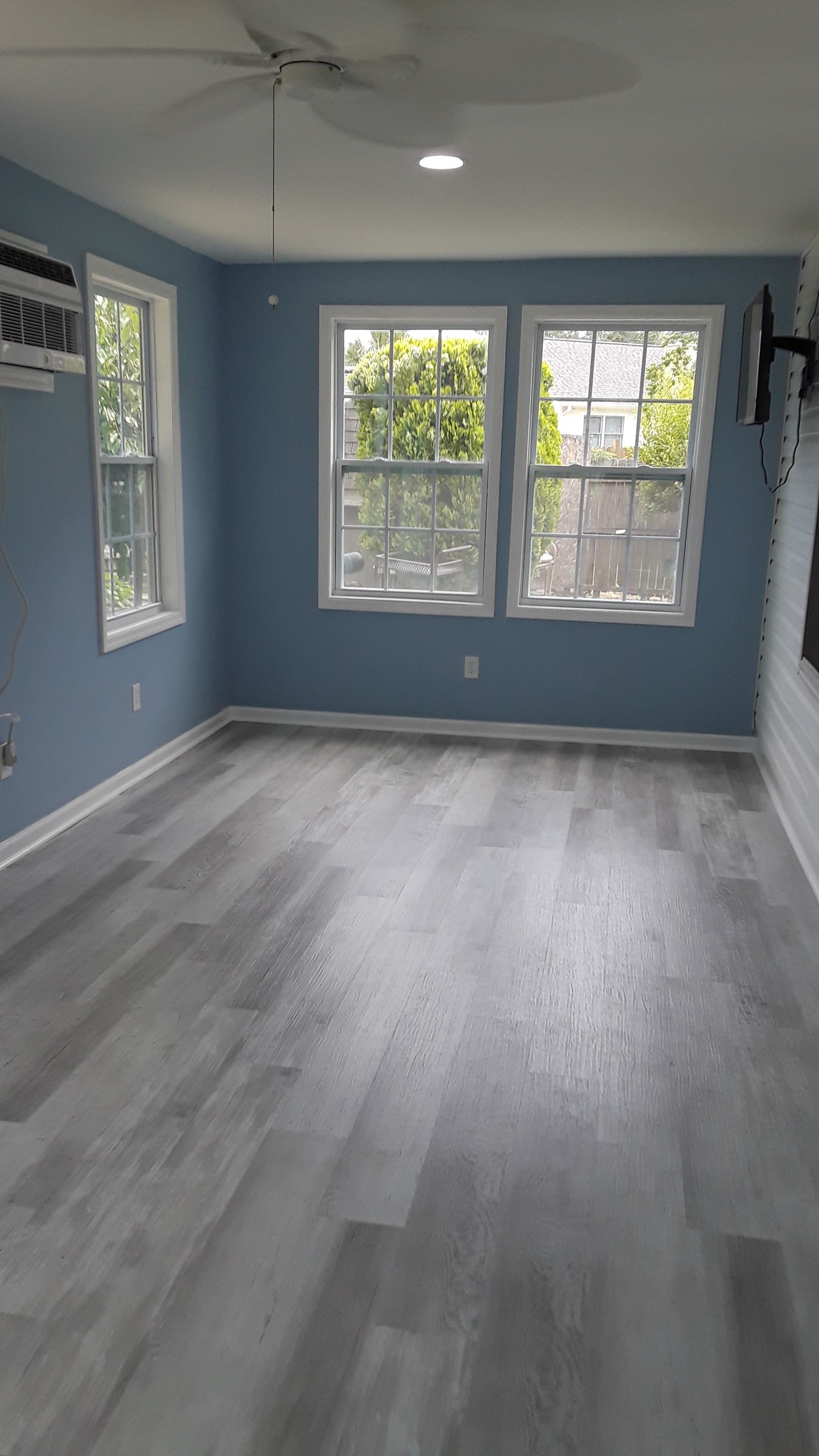 An empty room with blue walls, light grey wood-look flooring, two windows, and a ceiling fan.