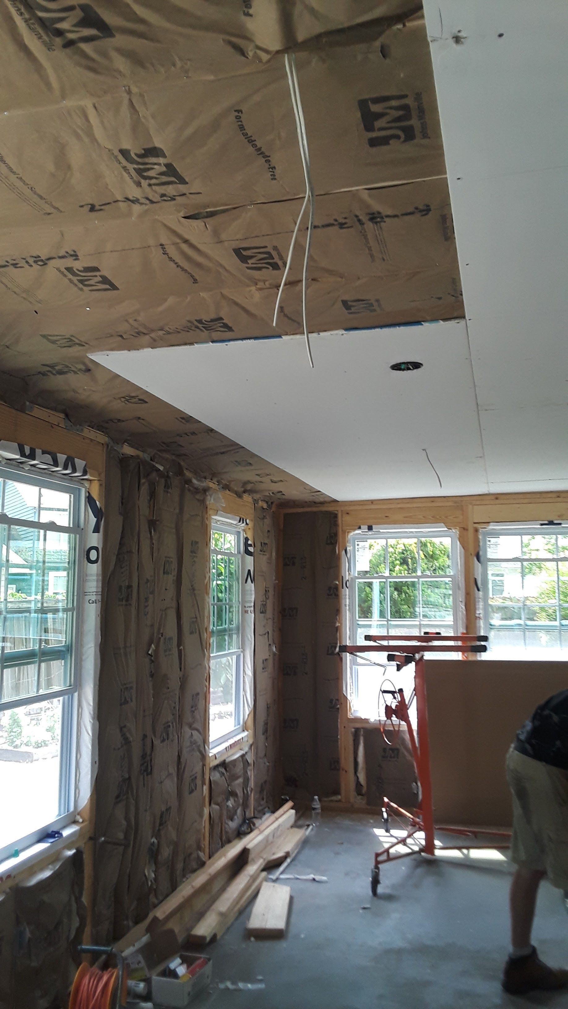 An interior room under construction with exposed wall insulation, partially installed drywall on ceiling, and two windows.