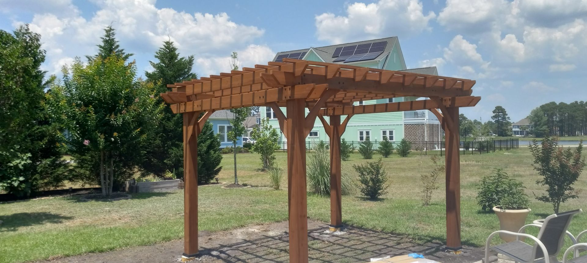 A wooden pergola stands in a grassy backyard with a partially constructed house in the background under a blue sky.