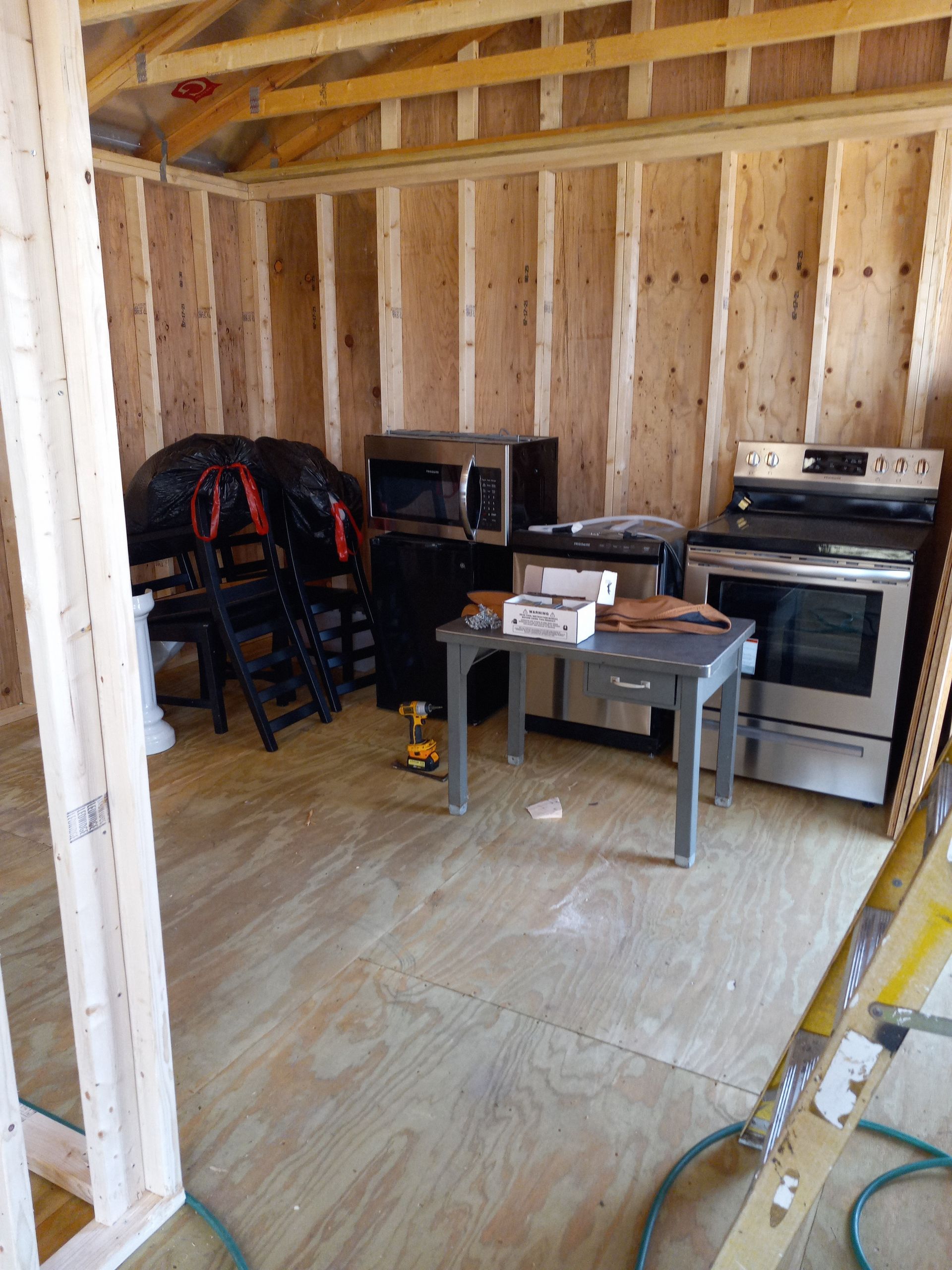 Interior of a wood-framed shed under construction with an oven, microwave, and small table placed on the plywood floor.
