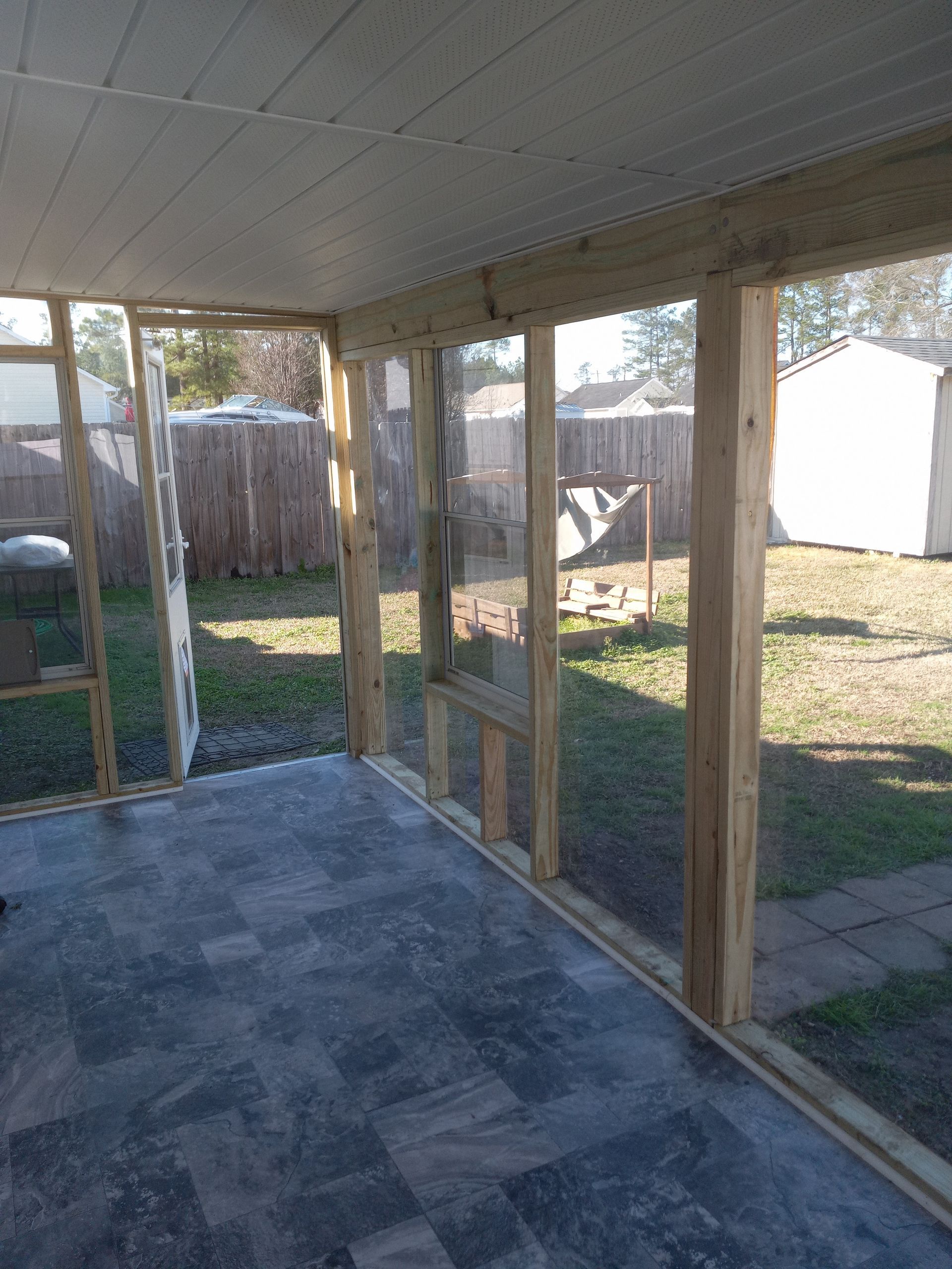 A screened-in porch with a gray tile floor, wooden support posts, and a view of a backyard with a fence and shed.