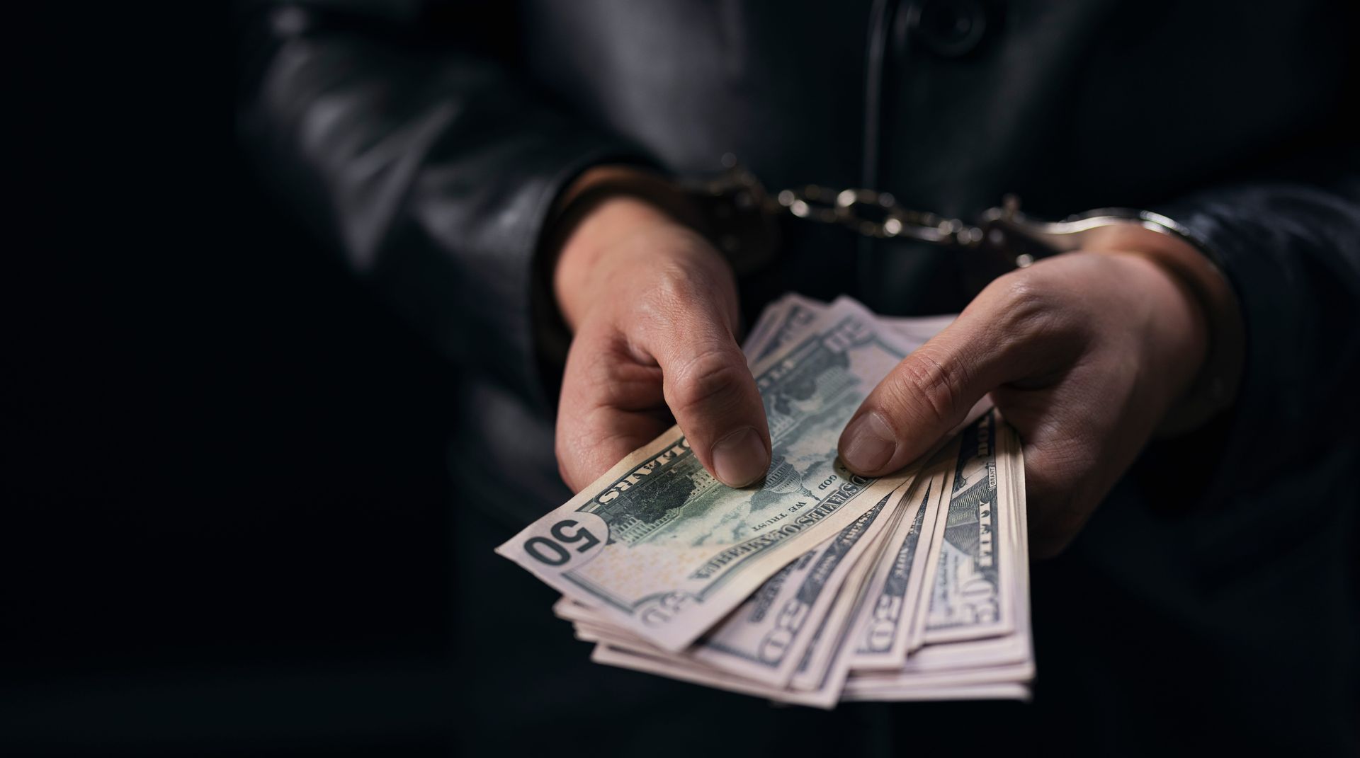 Hands counting a fan of U.S. dollar bills in dim light, with handcuffs visible on the wrists.