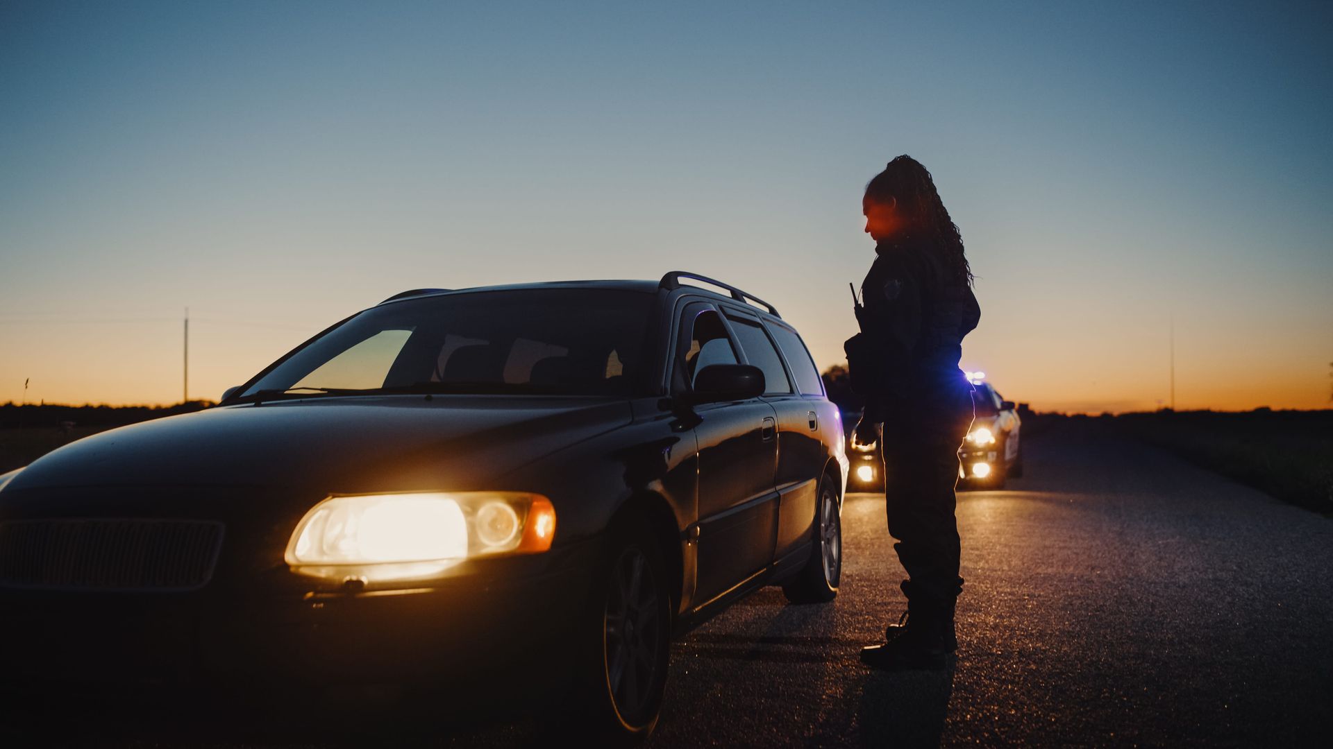 Car parked at dusk with headlights on; a person stands by the driver's door on a dark road.