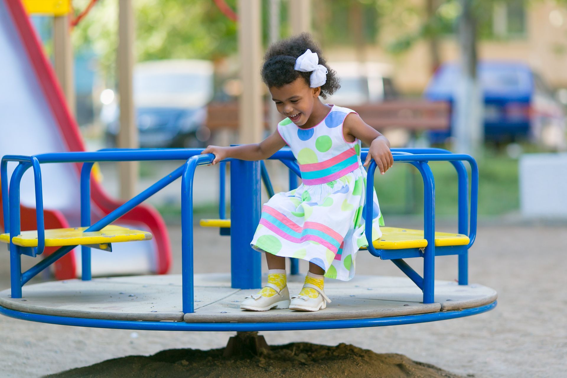 Girl in polka-dot dress on a blue and yellow playground merry-go-round, smiling at a park.