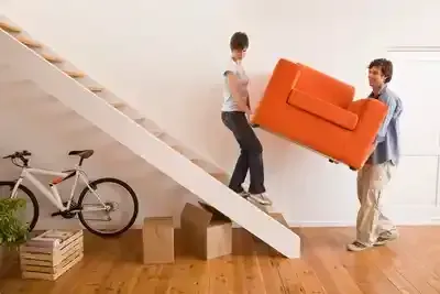 A man and a woman are carrying an orange chair up a set of stairs.
