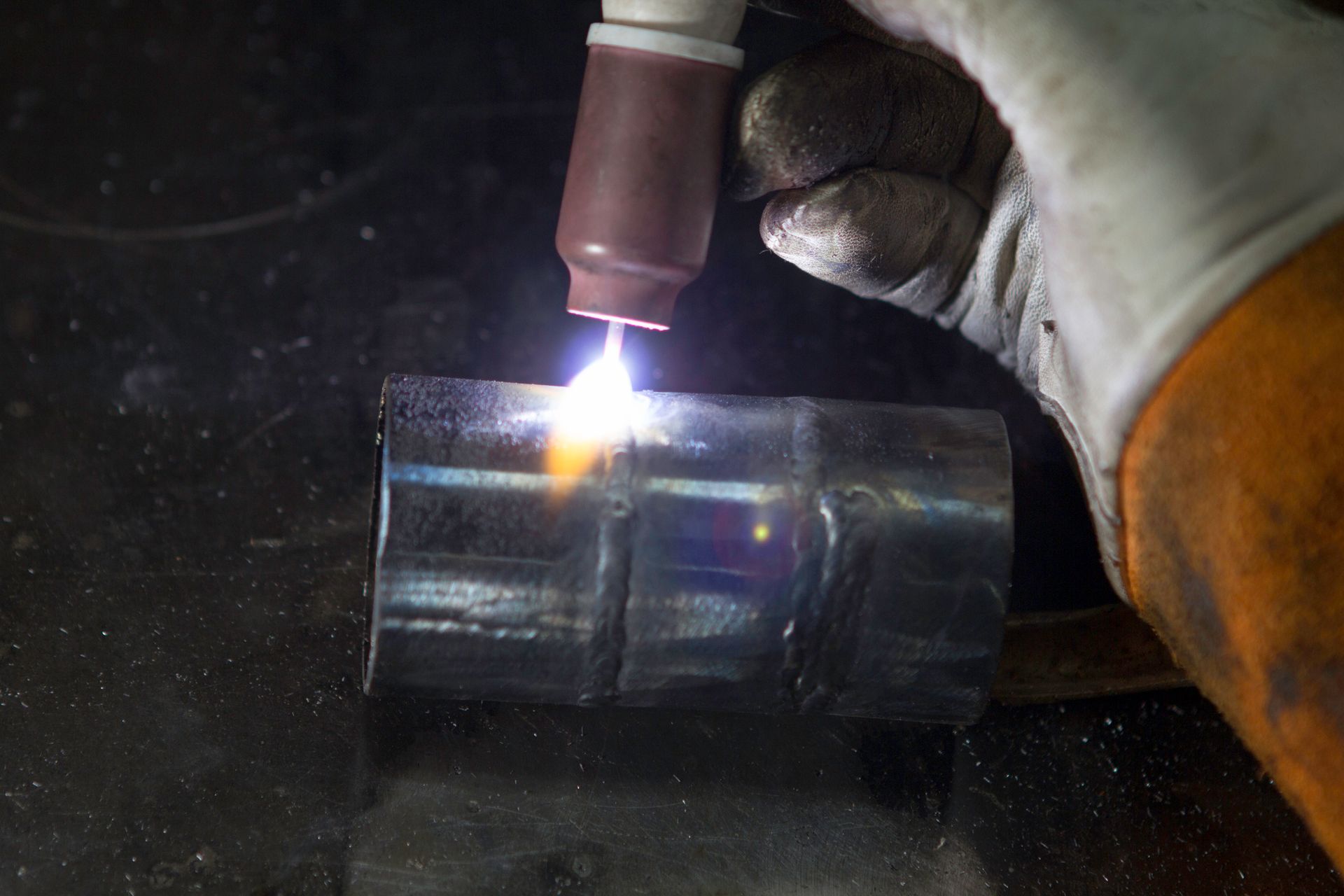 Welder using a torch to weld a metal cylinder. Bright sparks and a glowing orange flame visible.