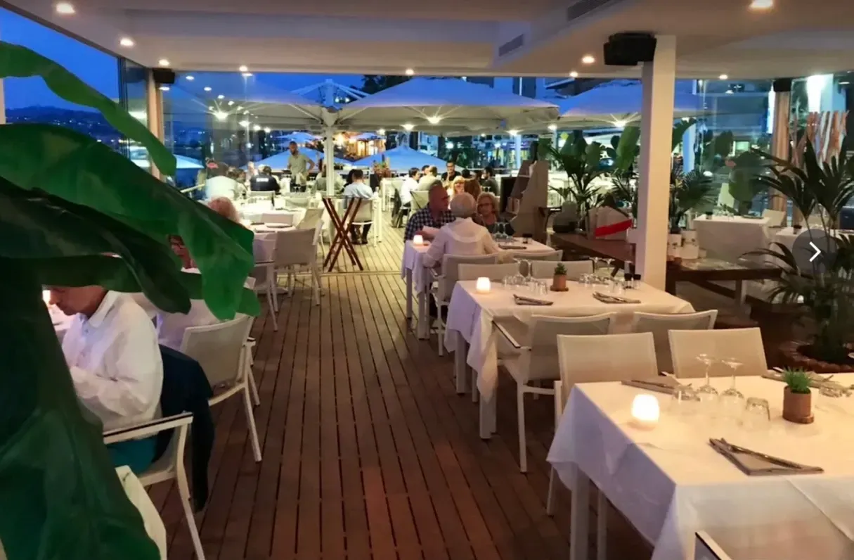 Restaurant en plein air le soir avec tables blanches, invités et parasols.