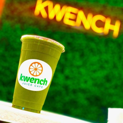 Green Kwench Juice Cafe cup with orange logo against a blurred green wall and Kwench sign