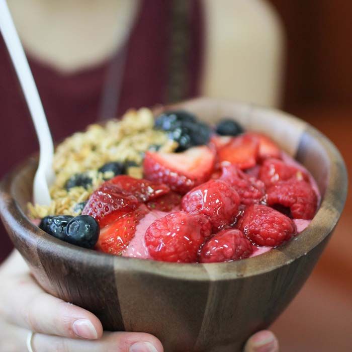 Wooden bowl of yogurt topped with strawberries, blueberries, and granola, held in hands with a spoon.