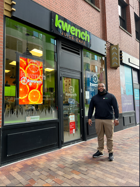 Man standing outside a Kwench storefront with bright green signage and an orange window display.